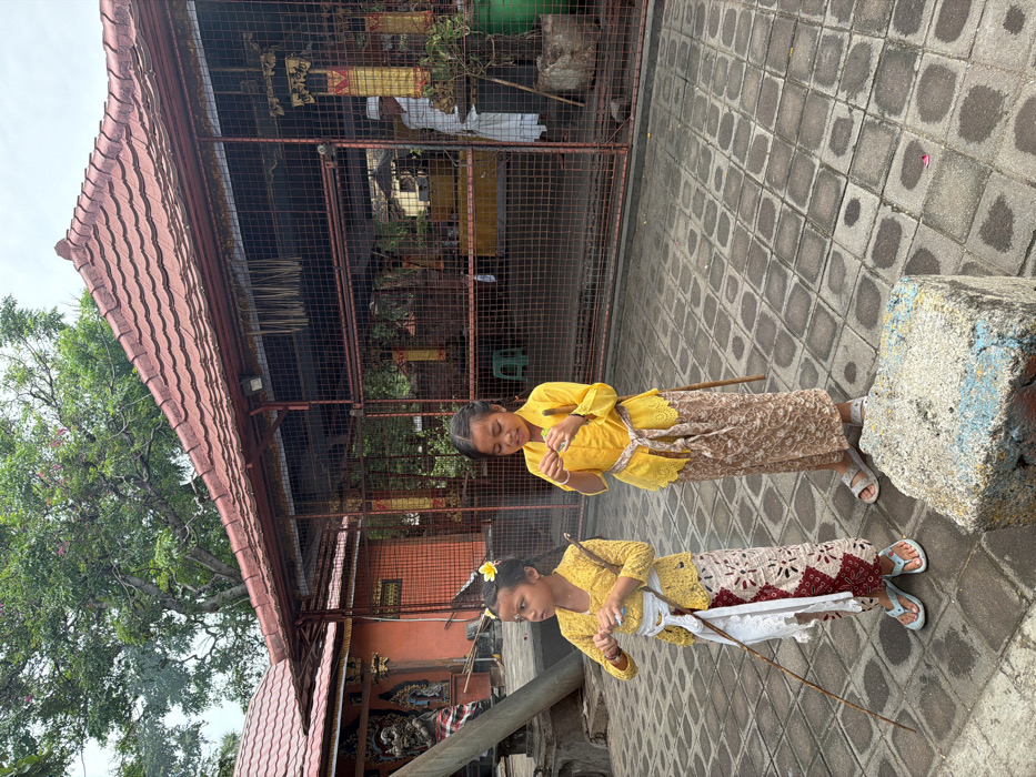 Two girls in yellow traditional clothing stand on a paved surface. The girl in the foreground has a white flower in her hair and holds a stick-like item. The other girl stands slightly behind. A building with a red-tiled roof and a metal enclosure behind them. Trees are visible on the left side, and a stone structure appears on the right edge.