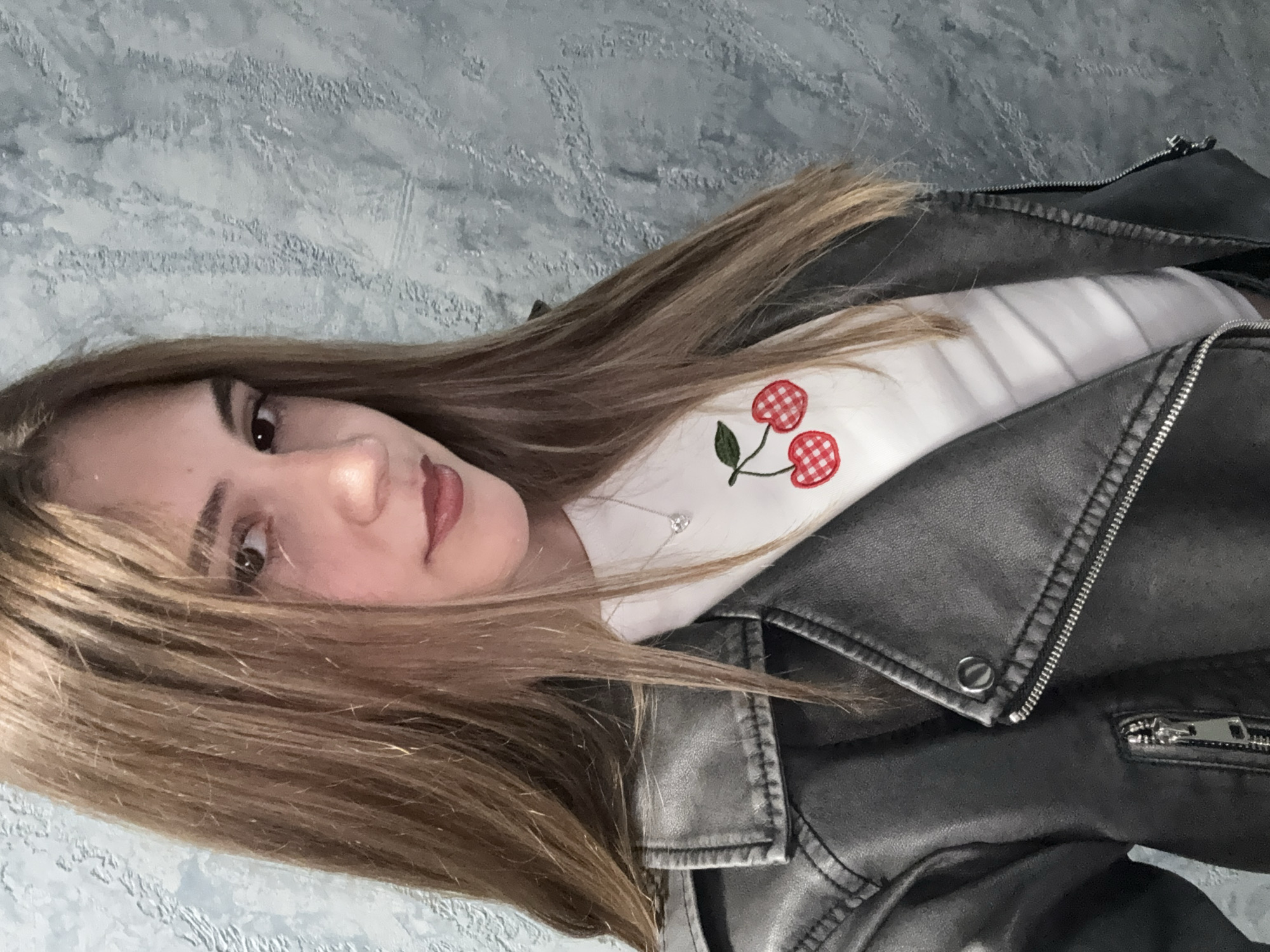 A young woman with long, brown hair is shown in a selfie. She is wearing a white, long-sleeved shirt with a cherry pattern on the front and a black leather jacket. A silver necklace is visible around her neck. The background appears to be a textured wall. The image does not show any information to determine a location in the city of Panagyurishte, Bulgaria.