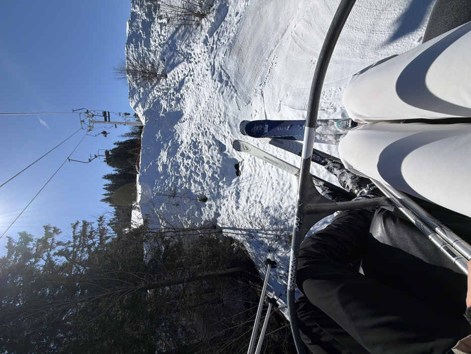 The image presents a first-person perspective from a ski lift chair overlooking a snow-covered ski slope in Manigod, France, on a clear, sunny day. In the immediate foreground, a metal safety bar, marked "WOODCORE 900," spans the lower frame. Two individuals are seated on the ski lift, their lower bodies and equipment visible. The person on the left wears black ski pants and holds two ski poles, with white and black Rossignol brand skis dangling below. The person on the right wears white ski pants, with blue and black Elite brand skis suspended beneath.
The ski slope descends directly below, showing packed and uneven snow with visible ski tracks. To the left, a dense forest of evergreen trees lines the slope, casting long shadows. Scattered bare deciduous trees are visible on the snowy incline. In the mid-distance, a metallic ski lift tower supports the overhead cable, along which several empty ski chairs are visible. The sky is bright blue, with a prominent sun flare in the upper left quadrant. Evergreen trees are also visible on a distant ridge line in the background.