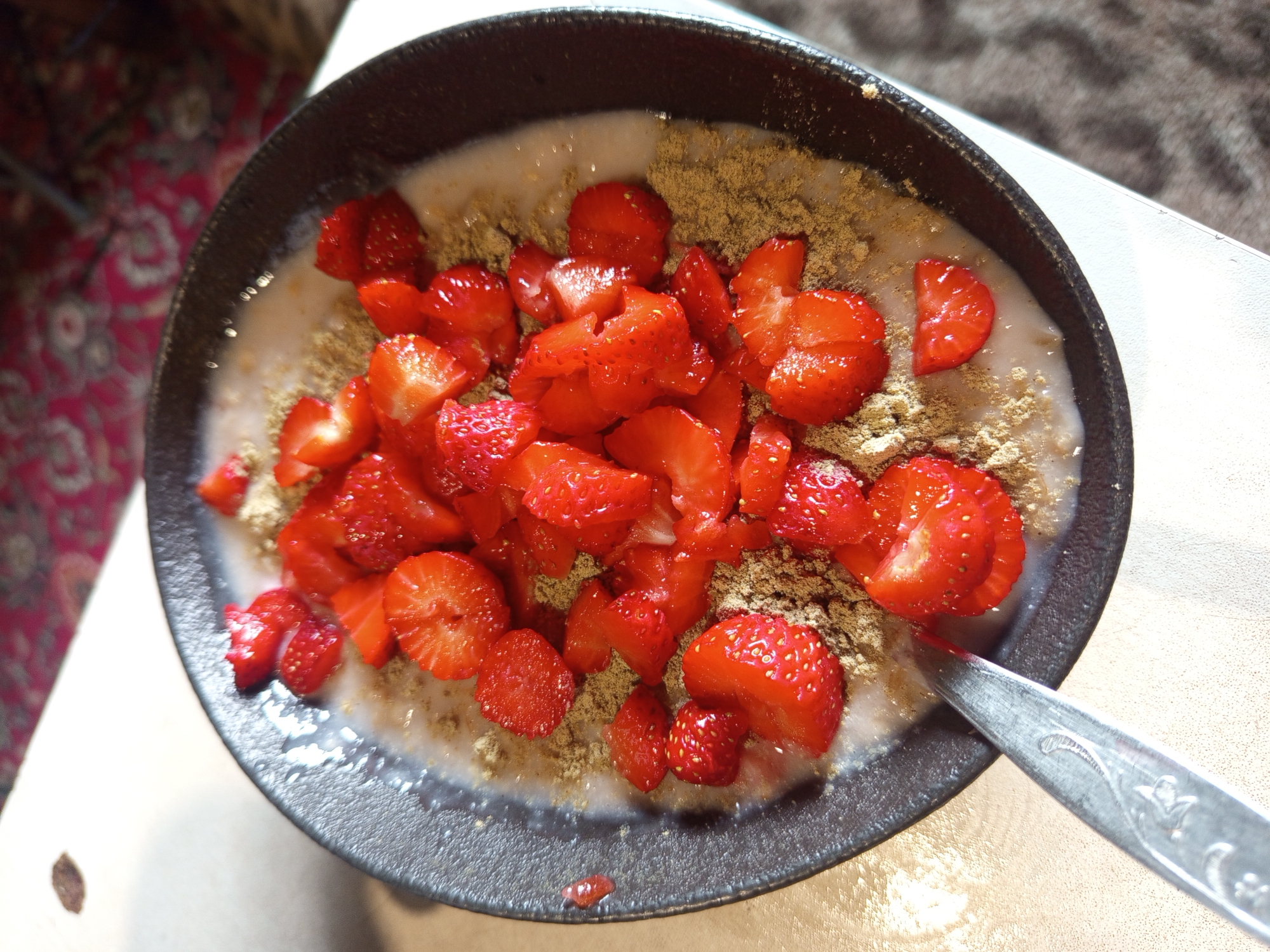 An overhead shot captures a dark-rimmed bowl filled with creamy oatmeal topped with sliced strawberries and a sprinkle of a light brown powder, possibly cinnamon or a spice blend. A silver spoon with an ornate handle rests partially submerged in the oatmeal, its tines pointing towards the left side of the frame. The bowl sits on a textured, light-colored surface, likely a table, with a blurred background showing hints of a patterned rug and a hint of a brown object. The image composition emphasizes the food, with the bowl occupying the majority of the frame. This image was taken on a table in a room in Ostrovtsy, Russia.