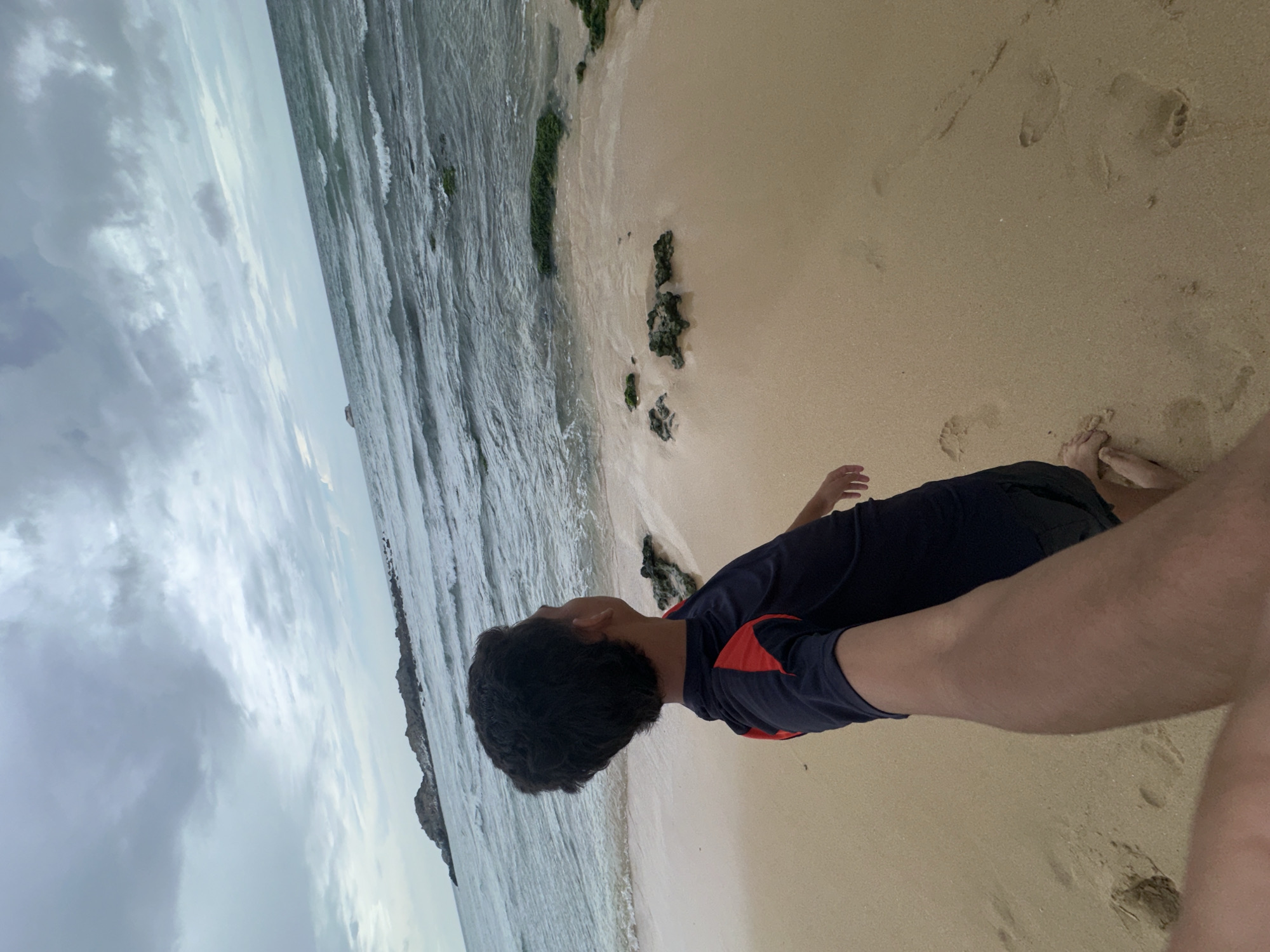 A boy stands on a sandy beach, facing away from the camera and looking out at the ocean. The sky is overcast with clouds. The ocean has waves and some rocky outcrops are visible in the distance. The sand has footprints on it. The boy is wearing a dark blue shirt with red accents on the shoulders and dark shorts. The image appears to be taken in Altagracia, Venezuela.