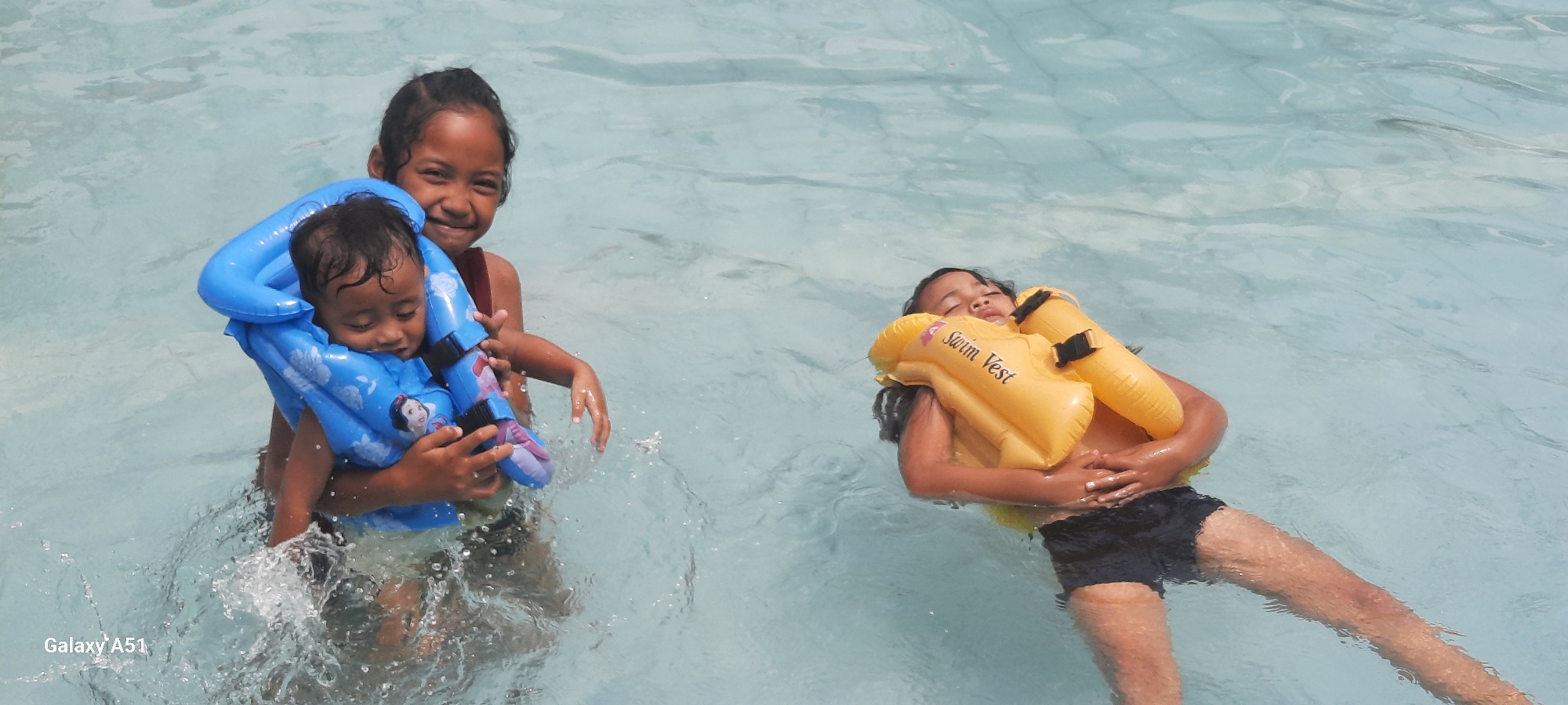 In the clear, blue waters of a swimming pool in Popoh, Indonesia, two young girls are enjoying a day of aquatic recreation. On the left, an older girl with dark hair pulled back, smiles widely as she embraces a baby wearing a bright blue inflatable swim vest adorned with images of Disney princesses. The baby, with dark, wet hair, is nestled securely in the older girl's arms, its face appearing content or sleepy. Water splashes around them, indicating movement and play. To the right, a second girl with dark hair floats on her back, wearing a yellow inflatable swim vest that reads "Swim Vest." She has her eyes closed and a relaxed expression, her body partially submerged in the tranquil pool. The pool's surface is clear with subtle light reflections and ripples, suggesting a bright, sunny day. The scene captures a moment of childhood joy and safety in a recreational setting.