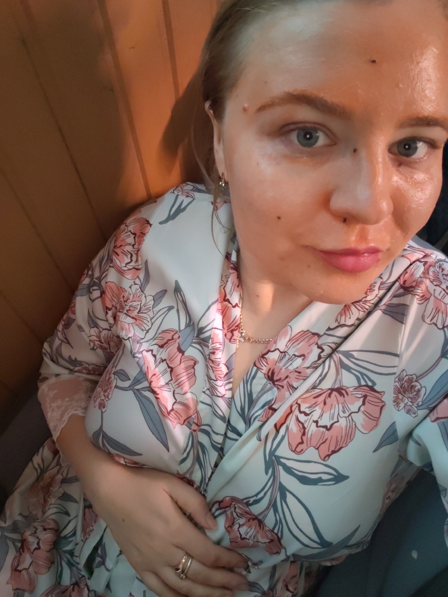 A fair-skinned woman with blue eyes and blonde hair is posing for a selfie indoors. She is wearing a white robe with a pink floral pattern and a delicate gold necklace. Her expression is subtle, with her lips slightly pursed. The background consists of vertical wooden panels and what appears to be part of a gray chair or cushion. There is no information that specifically indicates that the picture was taken in Izhevsk, Russia.