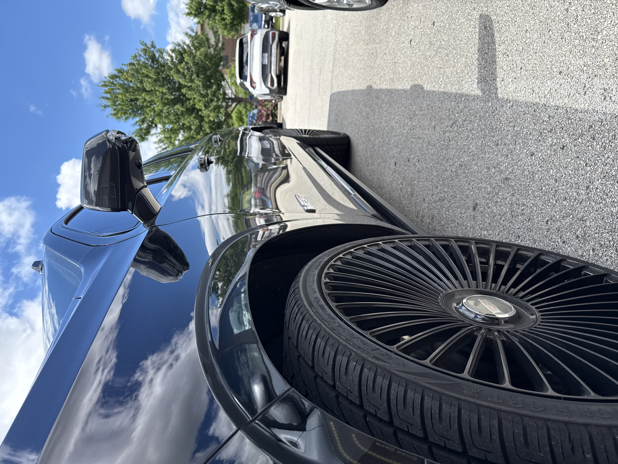 In Burr Ridge, Illinois, a black Chevrolet Tahoe, likely a 2021 or newer model, is parked in an asphalt lot under a bright blue sky with scattered clouds. The vehicle, viewed from a low angle and close-up perspective focusing on the front passenger side, exhibits a high-gloss finish reflecting the surrounding environment. The custom black multi-spoke rims are a prominent feature, fitted with 305/30ZR26 tires, suggesting a modified aesthetic. The vehicle's side mirror has a carbon fiber finish. In the background, other vehicles are visible, including a white Kia Sportage and a dark-colored SUV, as well as mature green trees. The scene appears to be a commercial area, possibly a retail parking lot. No people are visible in the frame.