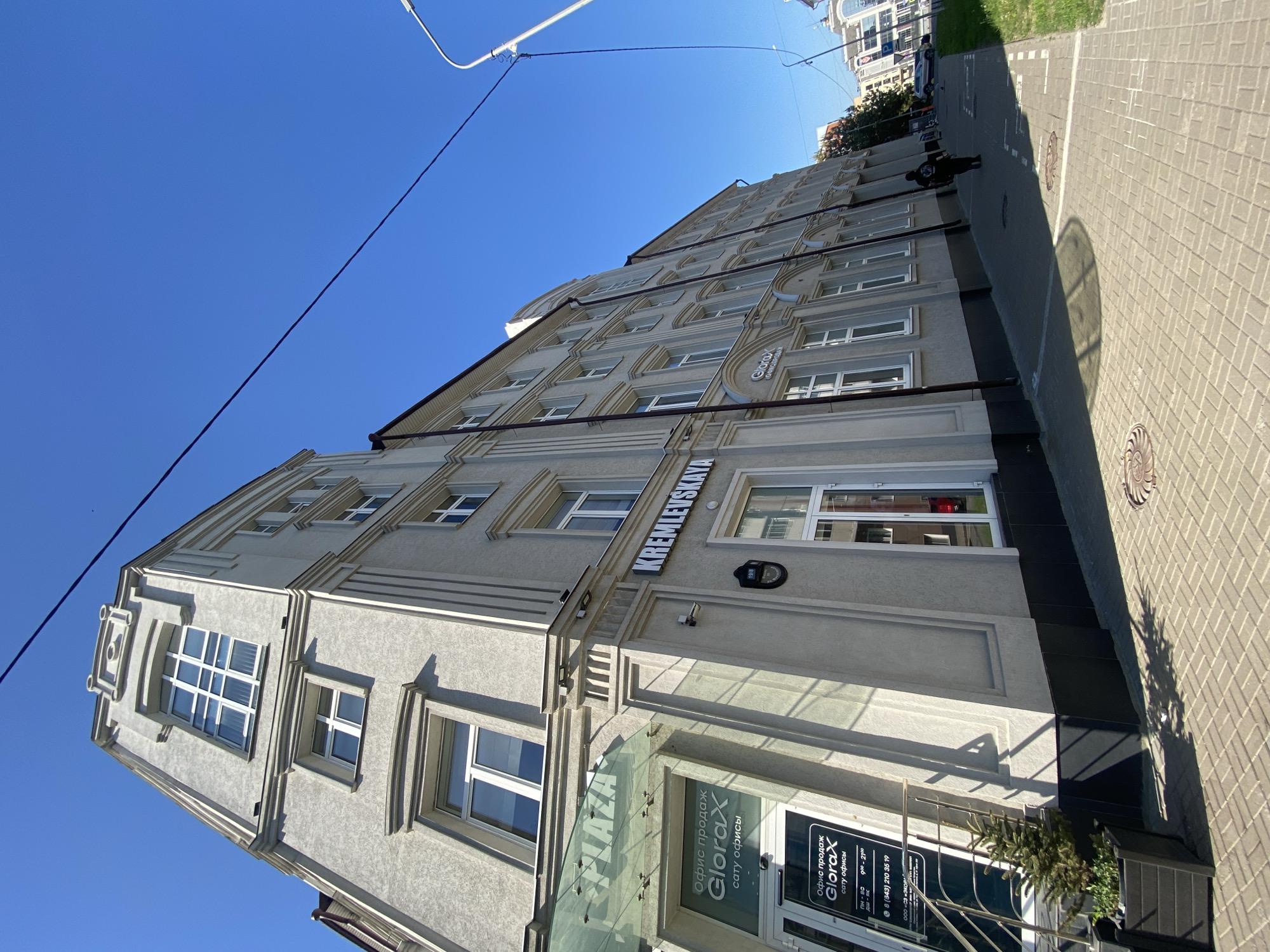 The image depicts a building in Kazan, Russia, identifiable by the "KREMLEVSKAYA" signage on the façade. The building is light-colored and features multiple windows arranged in rows. A sign on the entrance door reads "Glorax" and indicates the presence of an office. The street is paved with light grey blocks and a person can be seen walking alongside the building. A lamppost is visible overhead. The sky is clear and blue.