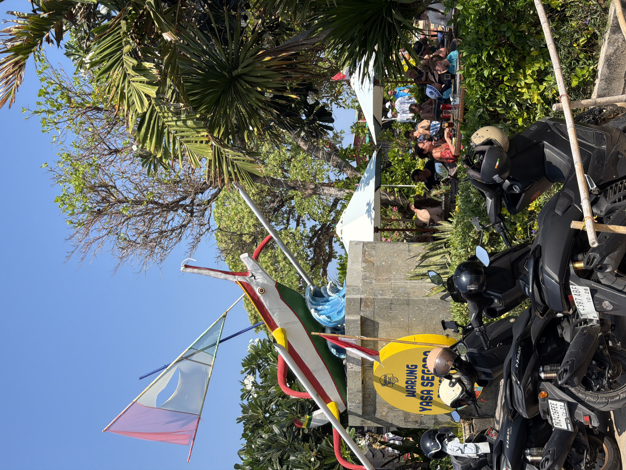 A sunny outdoor scene at a beachside restaurant features a decorated monument, several parked motorcycles, and patrons seated at tables. The monument is a stone structure topped with a colorful, stylized boat and a flag. A large yellow circular sign is mounted on the monument, reading "WARUNG YASA SEGARA" with a "Banana Boat" logo. Several black scooters are parked in the foreground, with helmets placed on their handlebars. In the background, people are seated at tables under umbrellas, some eating and drinking, while others converse. Lush green foliage and palm trees surround the area, with a clear blue sky overhead. The scene suggests a relaxed, tropical dining environment.