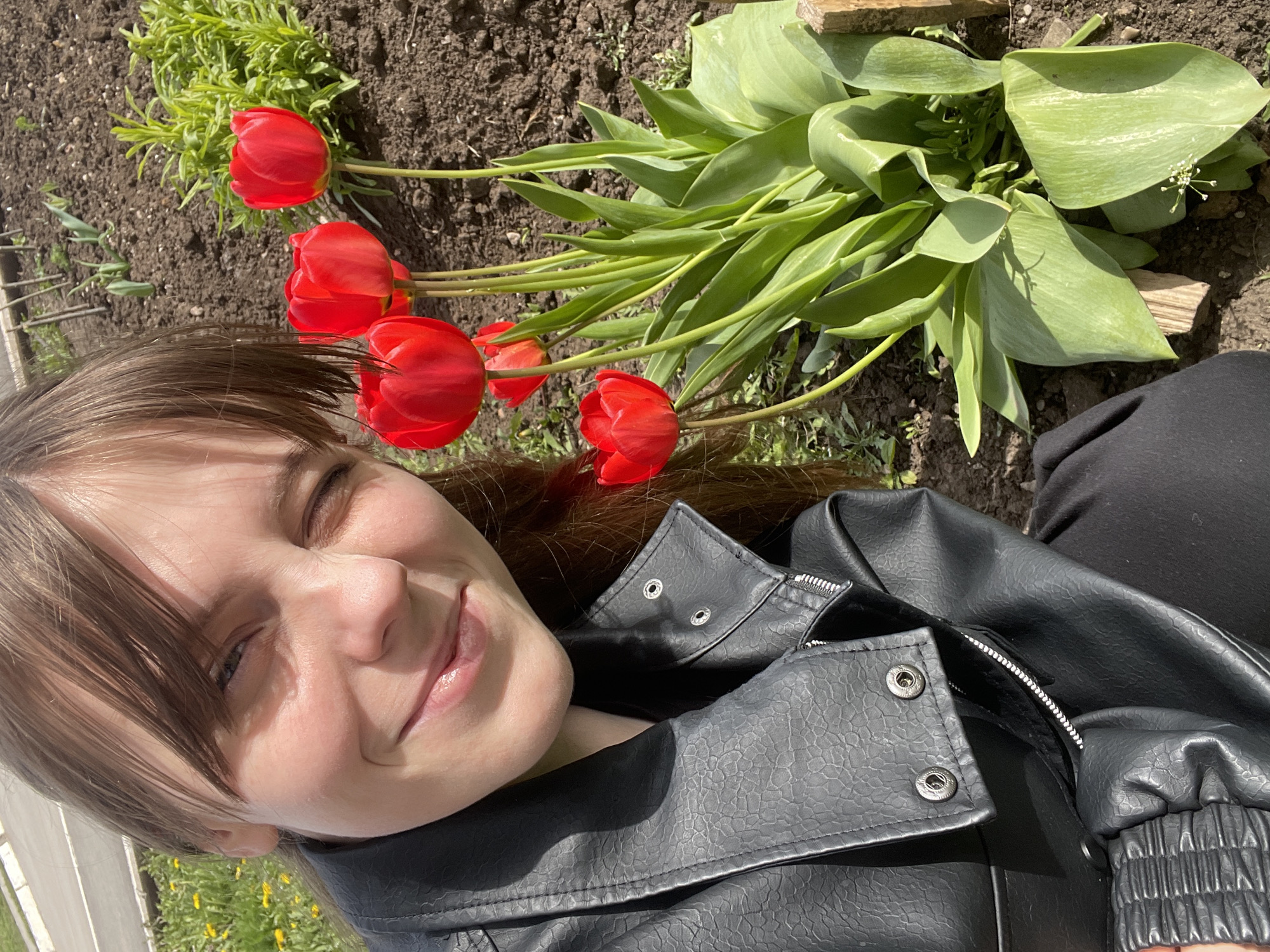 A young woman is lying down in a garden, next to a bunch of red tulips. She is wearing a black leather jacket and black pants. She is smiling and has her eyes closed. The background includes soil, grass, and other plants. The scene is outdoors. The location is not evident in the image. The city of Urussu, Russia, cannot be confirmed from this image.