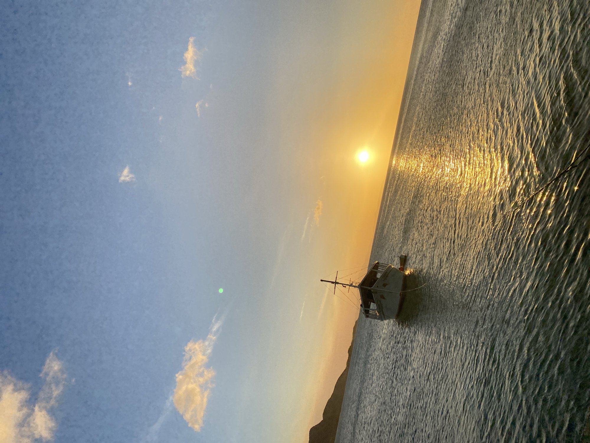 The image depicts a coastal scene in Juan Griego, Venezuela, at either sunrise or sunset. A large, light-colored fishing or utility boat, featuring an enclosed cabin and a mast with rigging, is anchored in the calm, rippling water. A smaller, dark-colored dinghy is tethered to the stern of the larger vessel. A rope from the main boat extends towards the bottom of the frame. The sun is positioned low on the horizon to the right, casting a prominent golden reflection across the water's surface, which extends from the horizon to the foreground. A dark, undulating landmass, appearing as hills or mountains, is silhouetted against the left horizon. The sky transitions from a light blue at the top to orange and yellow near the horizon, with scattered white and orange-tinged clouds. A green lens flare is visible in the upper central portion of the sky. No individuals are present in the scene. The image is captured at a slight upward tilt from left to right.