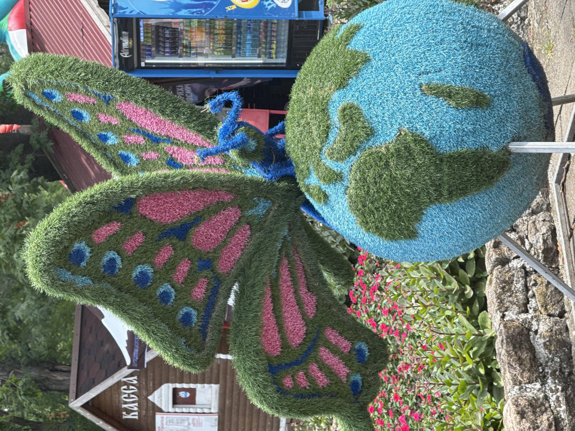 In the city of Yeysk, Russia, a garden sculpture depicts a large butterfly with intricate pink and blue wing patterns made of green, pink, and blue artificial grass. The butterfly is perched atop a spherical representation of the Earth, also crafted from artificial grass in blue for oceans and green for continents. A small blue insect figure, presumably the butterfly's body, is positioned on the Earth sculpture. Behind the sculpture, a wooden building with a dark roof and a sign reading "КАССА" (Cashier) is visible, along with a vending machine labeled "Adrenaline Rush". A bed of vibrant pink flowers and lush green plants occupies the mid-ground, separated from the sculpture by a low stone wall. The scene appears to be set outdoors in a park or public space on a clear day.