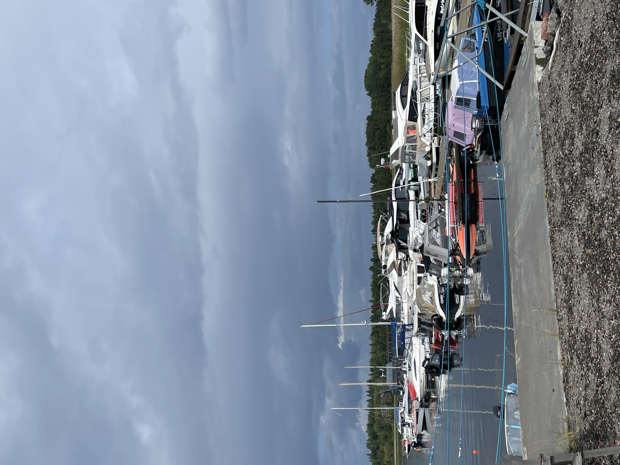 The image depicts a marina on an overcast day. Numerous boats of various sizes and types are docked in the water, including sailboats and motorboats. The boats are lined up along piers, with some secured by ropes. The water is dark and reflects the sky and the boats. In the background, a dense line of green trees forms the shoreline, with a field visible beyond the trees. The sky is filled with heavy, dark clouds, suggesting potential rain. The foreground shows a concrete pier with gravel on its surface. No people are visible in the image. The location is identified as Berozovo, Russia.
