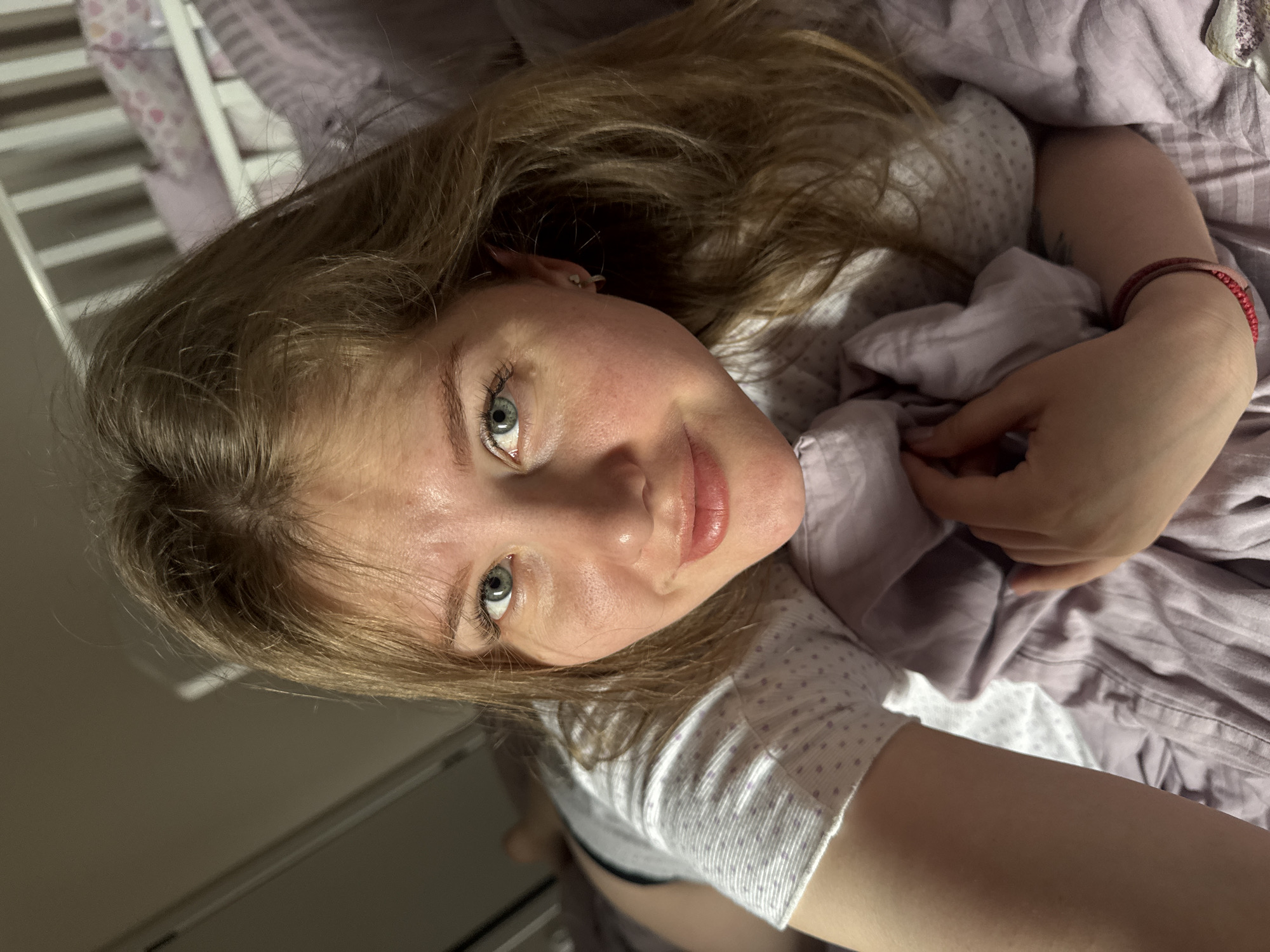 A young woman with fair skin, light brown hair, and blue-gray eyes is lying in a bed with lavender-colored sheets and pillowcases. She is wearing a white and gray polka-dotted shirt. Her right arm is visible, with a red bracelet on her wrist. The background shows part of a white crib or bed frame. The location is likely an indoor residential setting, given the bed and crib, but the specific details needed to confirm the city of Gornyi (Dvurechenskoe S/P), Russia are absent from the image.
