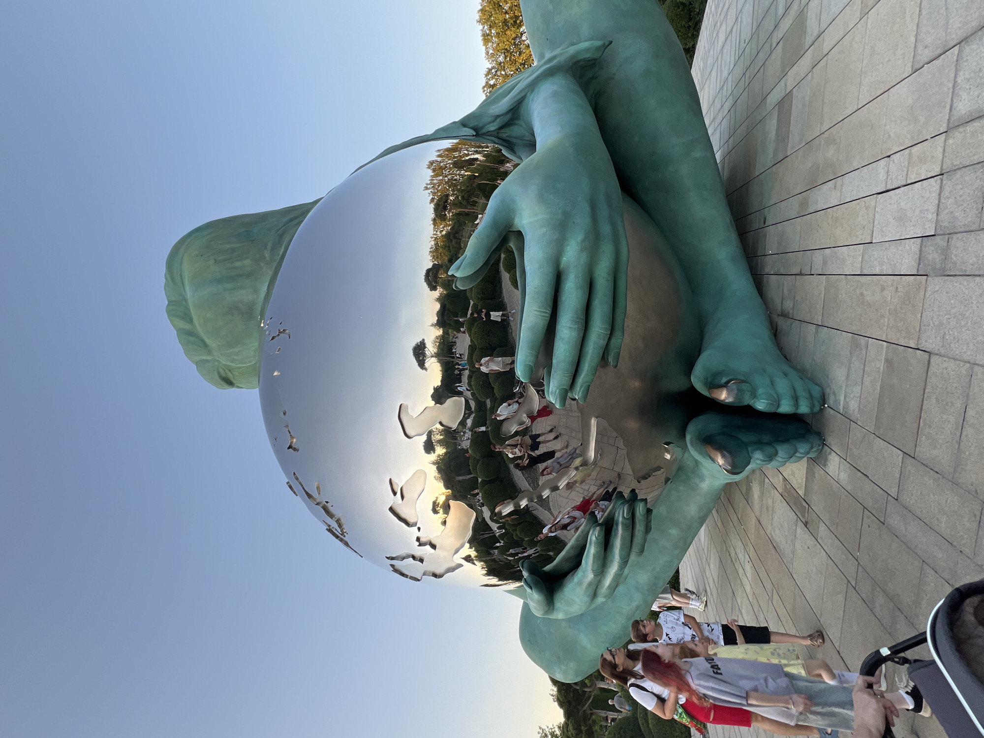 The image shows a large bronze sculpture of a reclining figure cradling a reflective globe. The sculpture is situated outdoors on a paved area surrounded by manicured bushes and trees. Several people, including adults and children, are observing the sculpture. The reflective globe mirrors the surrounding environment, showing the sky, trees, and people. This sculpture is located in Krasnodar, Russia, specifically in the "Gardens of Dreams" park, which is a popular attraction in the city. The scene appears to be during the late afternoon or early evening, with the warm light of the setting sun visible in the reflection.