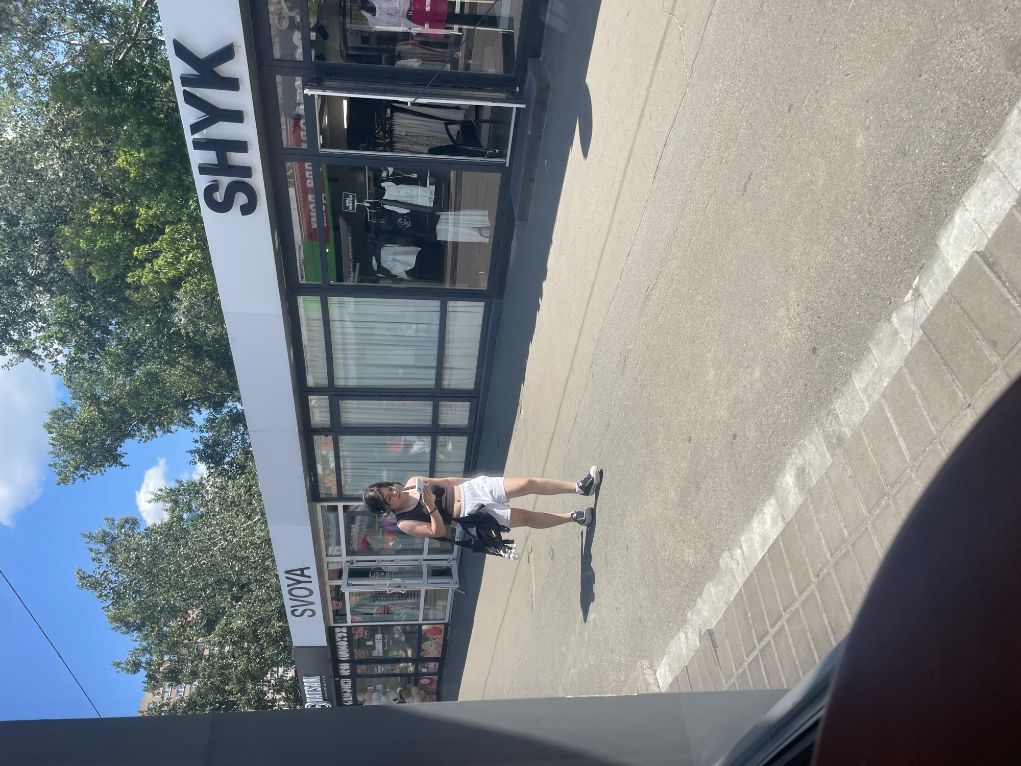 A young person stands on a sunlit sidewalk in front of a storefront, holding a phone and looking down. They are wearing a dark crop top, white shorts, and black and white sneakers. A black bag with white straps is slung over their left shoulder. The storefront has large glass windows displaying clothing on mannequins. Above the windows, prominent text reads "SHYK" in large black letters. Further down the storefront, the text "SVOYA" is visible. The scene appears to be a commercial street in Kyiv, Ukraine, bathed in bright sunlight, suggesting a warm, clear day. The shadows cast by the person and the building indicate it is likely midday. The surrounding environment includes trees with green foliage and a blue sky with scattered clouds. The sidewalk is paved with asphalt and bordered by a tiled curb. The overall atmosphere is casual and ordinary, depicting a moment of everyday life on a city street.