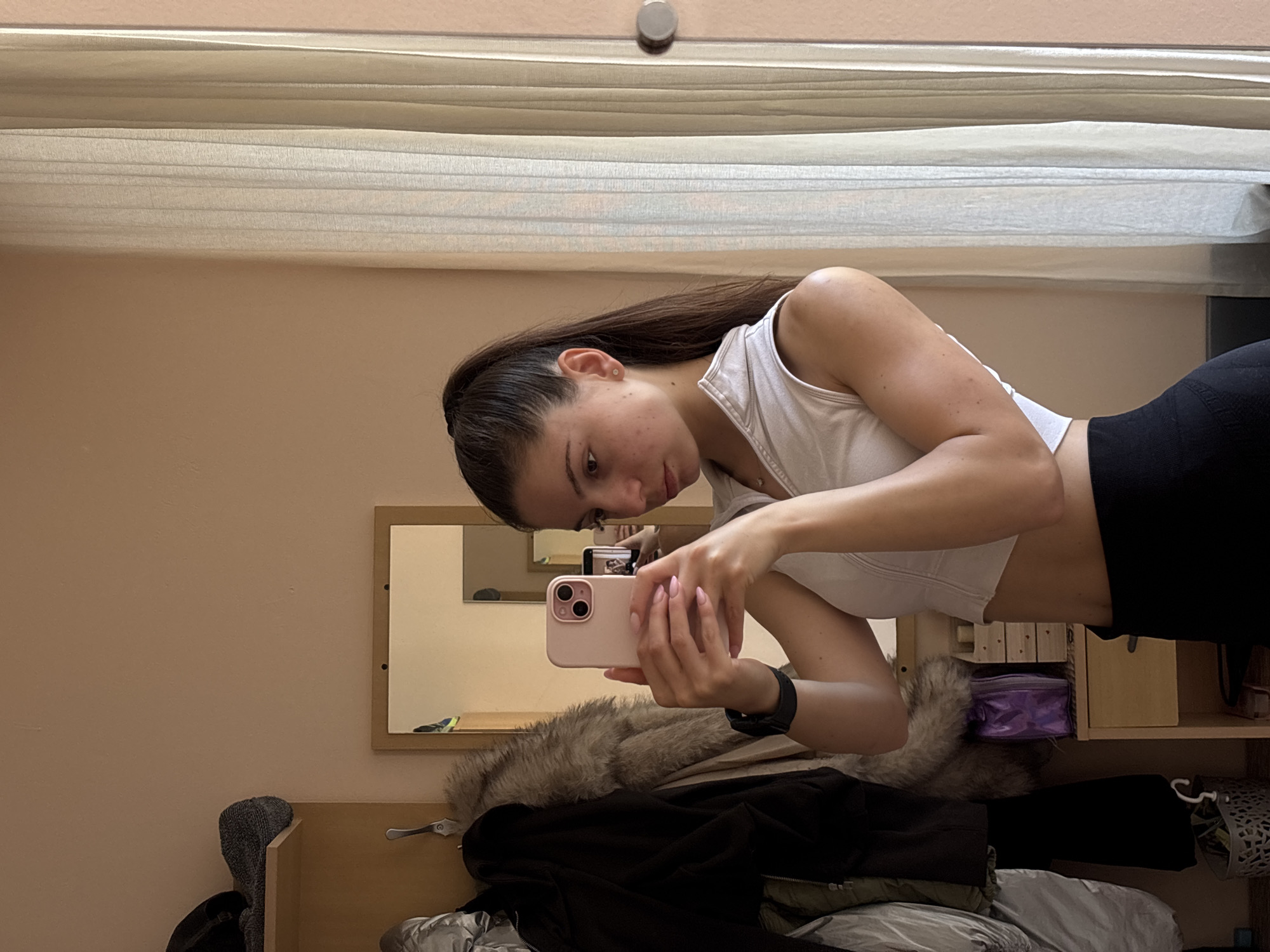 A young adult female is depicted taking a mirror selfie indoors, within what appears to be a residential or dormitory room in Varna, Bulgaria. She has dark brown hair styled in a high ponytail and is wearing a white sleeveless cropped top and black high-waisted athletic pants. A small, clear stud earring is visible in her left ear, and a delicate silver-colored necklace adorns her neck. A black smartwatch is present on her left wrist, and her fingernails are painted a light pink shade.
The subject holds a light pink smartphone, equipped with a dual-camera system, with both hands, directing its camera towards her reflection in the mirror. Her gaze is directed towards the phone's screen or her reflection within the primary mirror.
The room features plain, light peach-toned walls. To the left of the subject, an open wooden wardrobe unit contains a shelf with a dark knit hat and another dark garment. Multiple jackets hang from hooks below the shelf, including one with a prominent faux fur collar and a light grey puffer jacket. A small grey, decorative basket containing various items, including a white S-hook, rests on the floor beneath the hanging clothes. To the immediate right of the wardrobe, a small wooden nightstand features a purple toiletry bag/pouch and a multi-tiered wooden organizer on its surface.
The central feature is a large mirror framed in light brown wood. Reflected within this main mirror, further in the background, is a smaller, rectangular mirror, which in turn reflects a portion of the room, including what appears to be a window or door frame and a colorful object with blue and yellow sections. To the right of the main mirror, a window covered by vertical, sheer, light-colored blinds or curtains is present. A circular silver-toned door handle or knob is visible on the far right edge of the frame.
The lighting is soft and warm, suggesting natural light from the window supplemented by ambient indoor illumination.