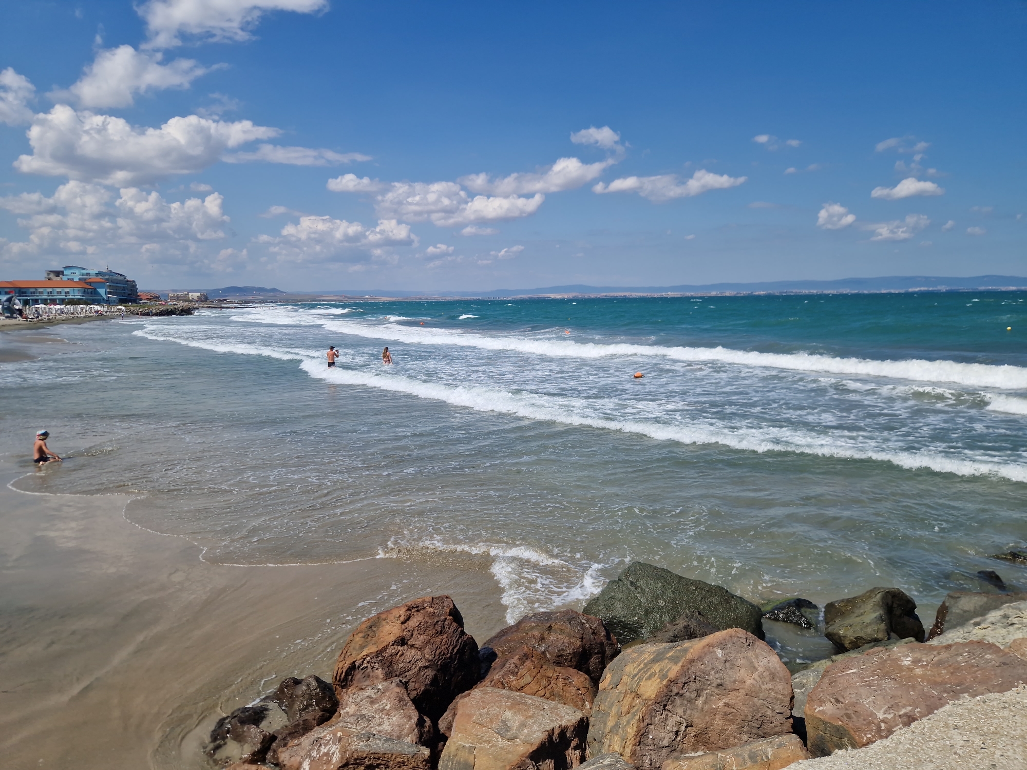 The image captures a sunny day at a beach in Pomorie, Bulgaria. The scene is dominated by the bright blue sky with scattered white clouds, meeting the turquoise ocean. Waves are rolling onto the sandy shore. In the foreground, large, reddish-brown rocks are visible along the edge of the water.
Further down the beach, a resort or hotel complex with blue and orange buildings is situated along the coastline. Several beach chairs and umbrellas are set up in front of the buildings.
In the water, a few people are visible. Two individuals appear to be standing or wading in the shallow water. Further out, a child is sitting in the shallow water on the left side of the frame. The sea has several buoys, some orange and one yellow, indicating areas for swimming or water sports. The overall impression is a pleasant summer day at a coastal resort.