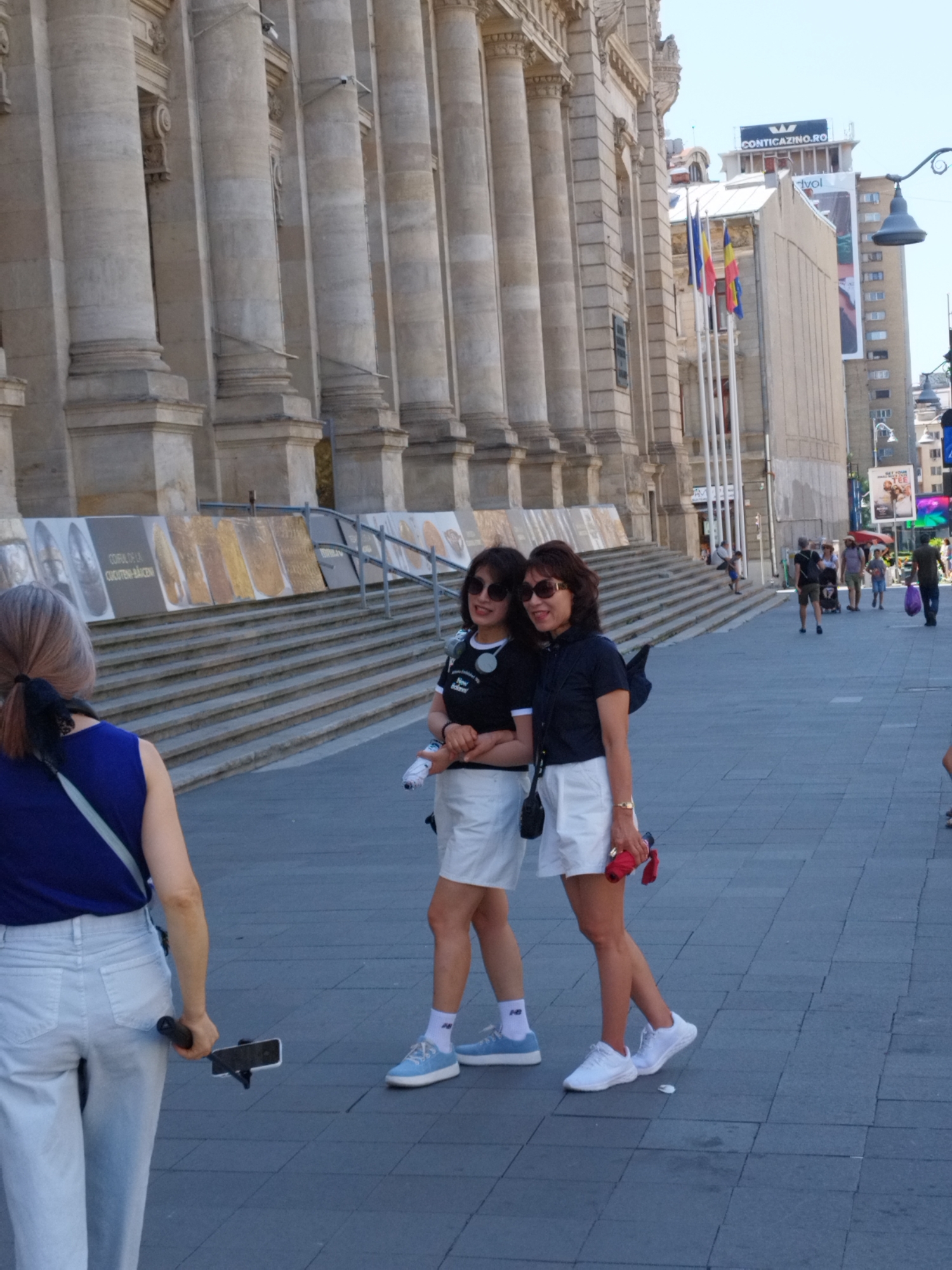 Two women, appearing to be of East Asian descent, are posing for a photograph in front of a large, classical-style building with tall columns. The woman on the left is wearing a black t-shirt with white lettering and white shorts, and light blue sneakers. The woman on the right is wearing a dark blue short-sleeved shirt and white shorts, and white sneakers. She is holding a small red umbrella in her right hand. Both women are wearing sunglasses. The woman on the left is holding a selfie stick with a phone attached. In the background, there are stairs leading up to the building, and several other people are visible walking in the distance. The building appears to be the National Museum of Art of Romania, located in Bucharest, Romania. The architecture is characteristic of buildings in the city's historical center.