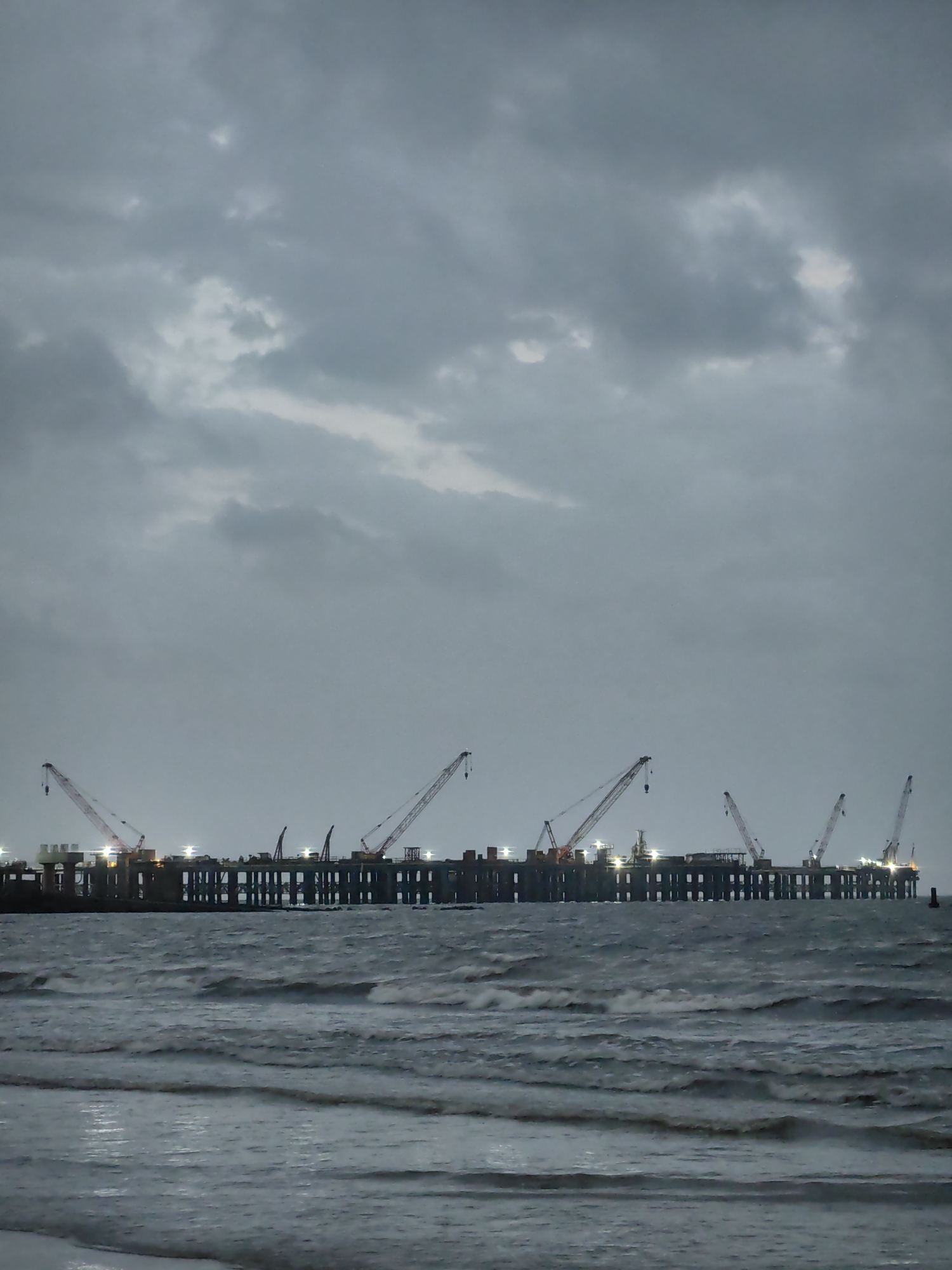 The image depicts a construction site on a pier or offshore platform under a cloudy sky, with waves in the foreground. The structure is supported by pilings and features several tall cranes, some of which are illuminated by lights. There appear to be some structures and equipment on the platform itself, and possibly a few indistinct figures. The overall scene suggests ongoing construction work, possibly at night or during overcast conditions. This is likely the Mumbai Trans Harbour Sea Link (MTHL) project, an under-construction bridge that will connect Sewri in Mumbai to Nhava Sheva in Uran, Navi Mumbai.