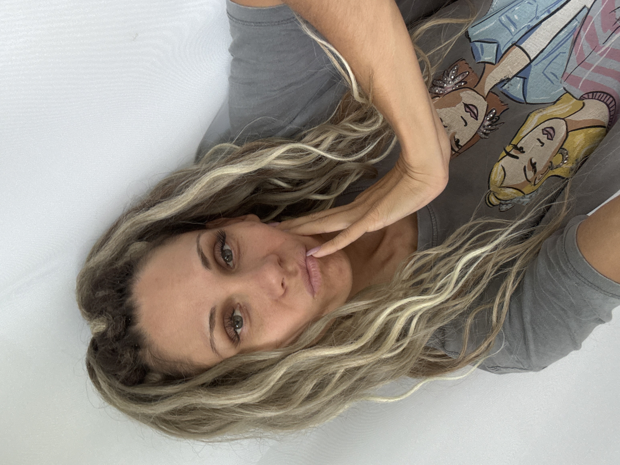 A woman with long, blonde, and brown hair tied up poses for a photo indoors against a white wall. She is wearing a gray t-shirt with a graphic of two cartoon women. Her right hand is touching her face as she pouts her lips. The woman has green eyes and is wearing makeup, including false eyelashes.