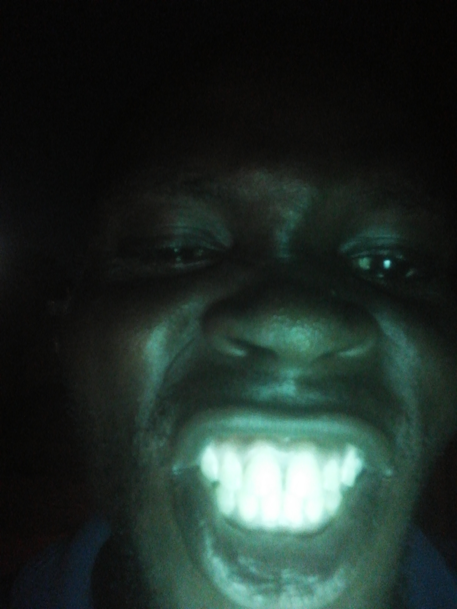 A close-up, dimly lit image of a person's face, dominated by a wide smile revealing glowing teeth. The person appears to have dark skin, with features visible despite the low light. The background is obscured by darkness, rendering the location unknown.