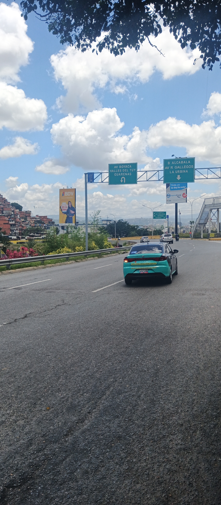 The image depicts a view from a vehicle on a multi-lane highway under a bright, cloudy sky. In the foreground, the asphalt road shows some wear and tear, with white lane markings. A teal-colored sedan is in the right lane, its rear end facing away from the camera. The car has a license plate that reads "AH 43408" and two yellow stickers on its rear bumper, one with text. Further ahead on the right side of the highway, a pedestrian overpass with metal stairs and railings is visible.
Overhead, two large green directional signs are mounted on poles. The left sign indicates "AV BOYACA VALLES DEL TUY GUARENAS" with an arrow pointing to the left and a U-turn symbol. The right sign directs to "B ALCABALA AV R GALLEGOS LA URBINA" with an arrow pointing straight and a right turn symbol. Below these signs, a smaller sign with the text "Seguros Venezuela" is visible. To the left of the highway, there is a billboard advertising "BDV" with an image of a woman holding a product. Beyond the billboard, a densely populated hillside with many brick and concrete structures is visible, characteristic of a barrio or favela. Vegetation, including trees and some flowering plants with pink and yellow foliage, lines the side of the road. The environment appears to be an urban setting in a warm climate.