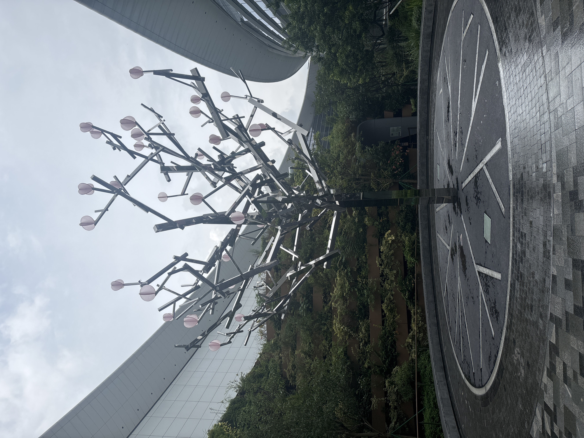 The image depicts a modern outdoor plaza with a large, abstract metal sculpture resembling a tree. The sculpture is composed of polished metal branches adorned with pink circular elements, possibly representing leaves or blossoms. It is centrally located within a circular paved area with white linear markings, suggestive of a design or pathway. The ground is wet, indicating recent rain.
Surrounding the plaza are several architectural structures and landscaping. To the left, a multi-tiered green wall covered in lush vegetation rises towards a sleek, modern building with a reflective glass facade and a curved, white roofline. To the right, another similarly designed building with a curved roof and glass windows is visible, partially obscured by trees. The sky above is overcast with diffused light.
The scene is devoid of people. The overall impression is one of contemporary urban design, emphasizing geometric forms, reflective surfaces, and integration of nature with architecture. This scene is located in Baiyun District, China.