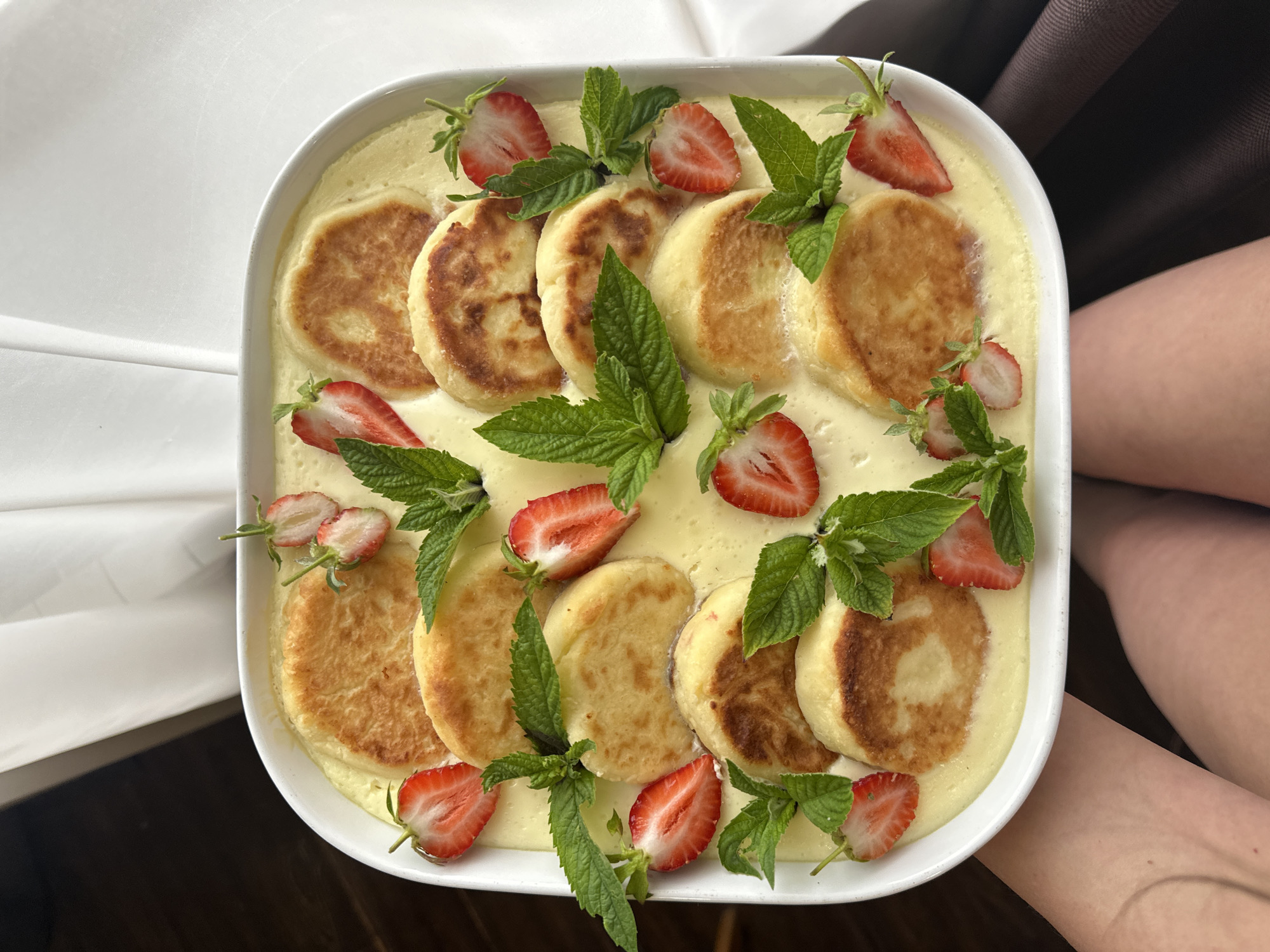 A white square dish filled with syrniki, a type of sweet cheese fritter, bathed in a creamy yellow sauce is held by a person in the foreground. The syrniki are arranged in rows, with several strawberry halves and fresh mint leaves scattered amongst them. The dish is placed on what appears to be a dark brown textured surface, possibly a tablecloth or clothing. The background consists of soft, blurred white fabric, suggesting a curtain or backdrop. The lighting is natural and bright, casting subtle shadows. The scene appears to be indoors, with no specific indicators pointing to the city of Chaikovsky, Russia.