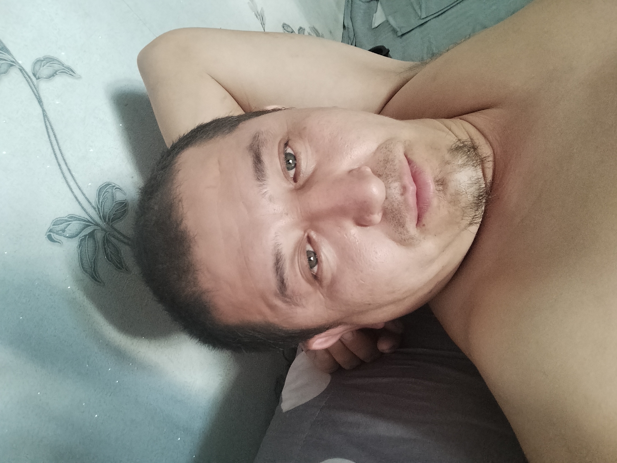 The image is a close-up, overhead shot of a man lying on a bed, with his head turned towards the camera. He has short, dark hair and stubble on his chin. His eyes are open, and he is looking directly at the camera. The man is shirtless, and his arm is resting behind his head. The background appears to be a bed with a patterned sheet and a pillow. The lighting is soft and natural. There is no indication of the city of Manas, Kyrgyzstan, or any urban environment in the image.
