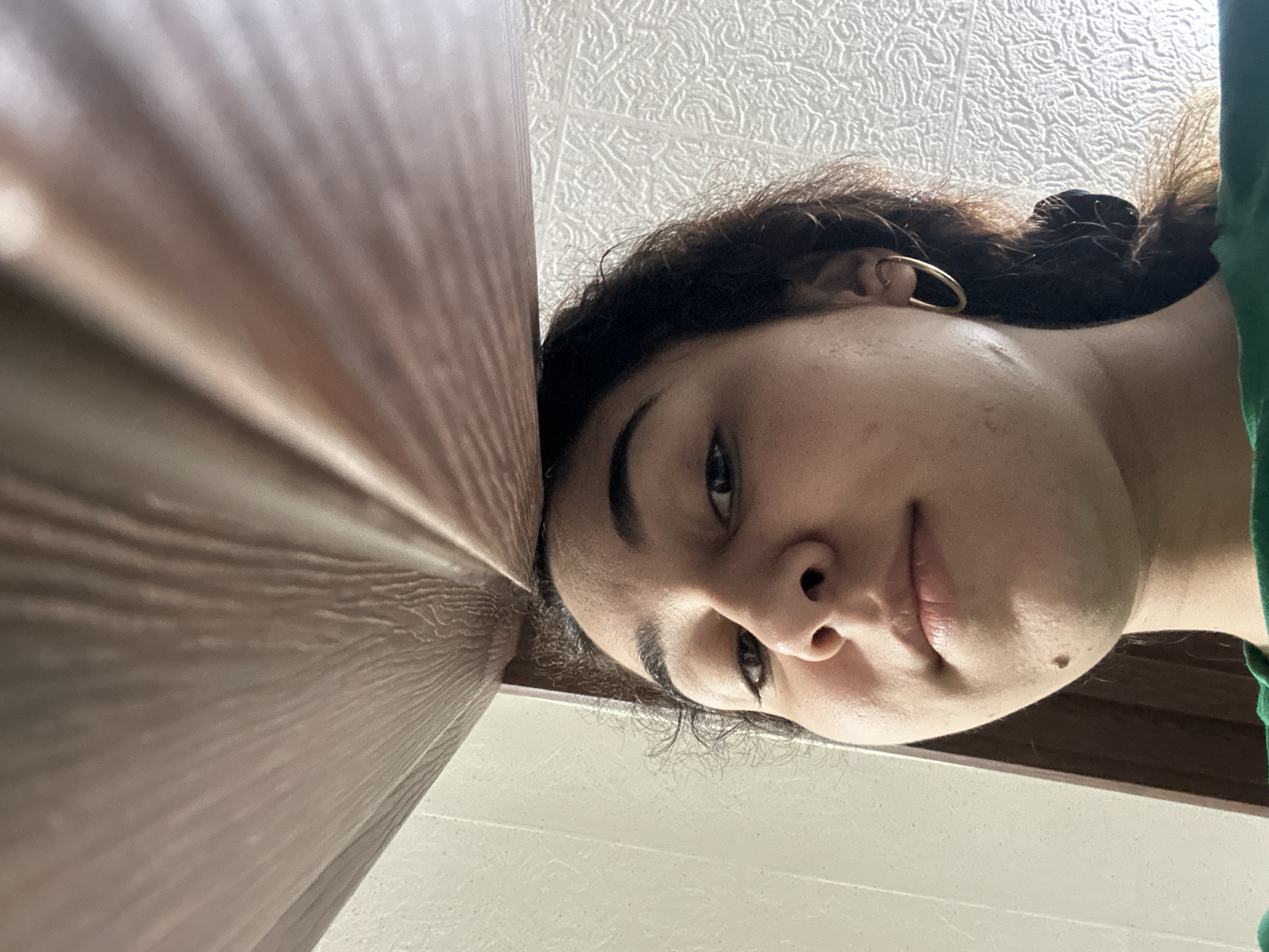 A close-up, slightly rotated view captures a person's face from a low angle. The person has fair skin, dark hair, and distinct eyebrows. A gold hoop earring is visible in the right ear. The person's face is close to a textured, brown surface, possibly a wooden door or wall. The background shows a textured white ceiling and a white wall. The environment appears to be indoors. The specific location in Burgas, Bulgaria cannot be determined from the visual information provided.
