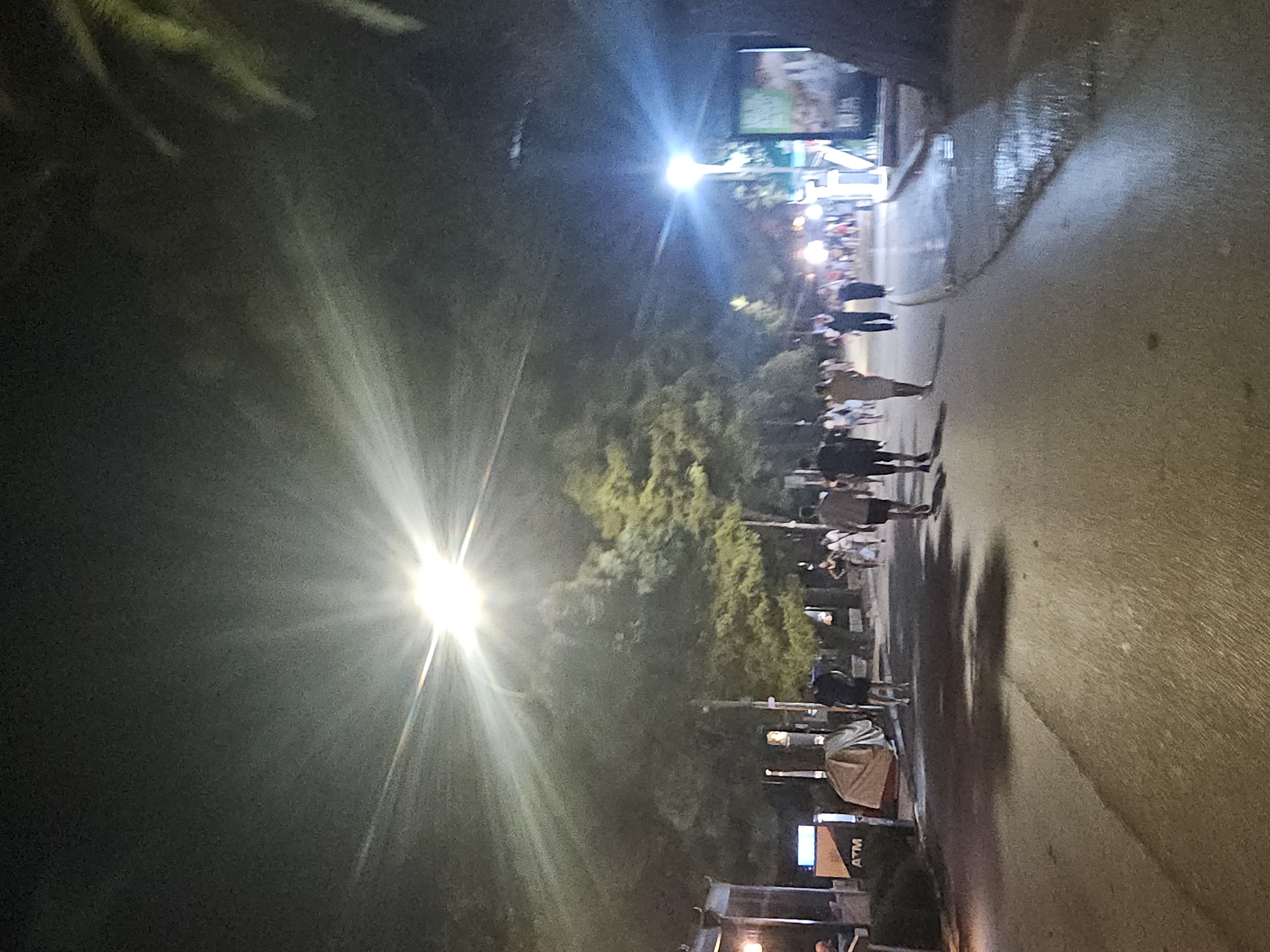 The image captures a nighttime scene on a paved walkway in Budva, Montenegro. The walkway is lined with trees, casting shadows on the ground. Several people are walking along the path, appearing to be engaged in casual strolling. A bright streetlamp illuminates the scene from above, creating a starburst effect and casting a strong glow. On the left side of the walkway, there's an ATM machine. The overall atmosphere suggests a public area, possibly a promenade or a street in a town, during the evening hours.