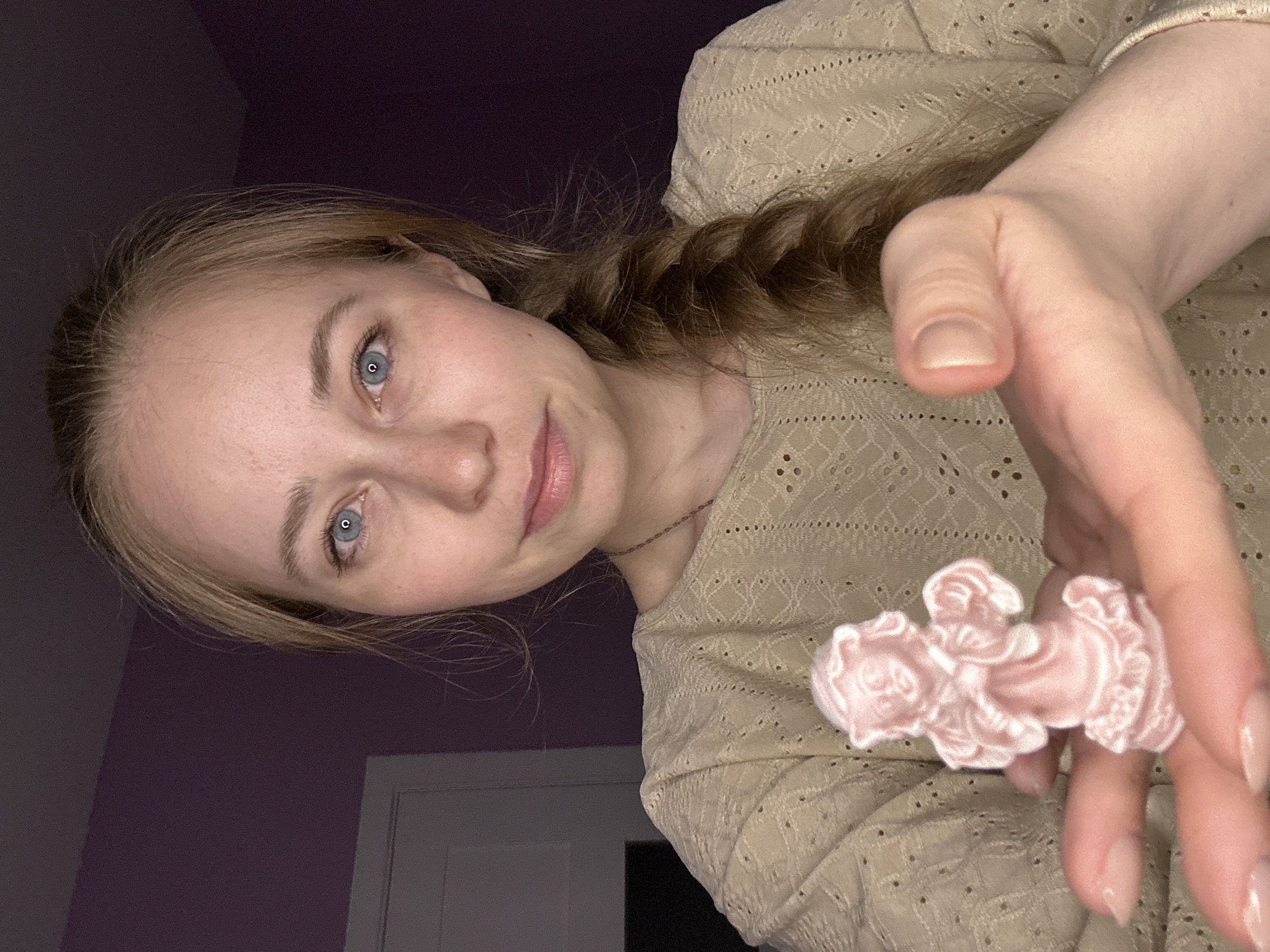 A woman with light blonde hair braided to her right is holding a small pink satin angel figurine between her index and middle fingers. She is wearing a beige lace-patterned top. The background consists of a partially visible white door and a lavender-colored wall. There is no definitive indication of Saint Petersburg, Russia.