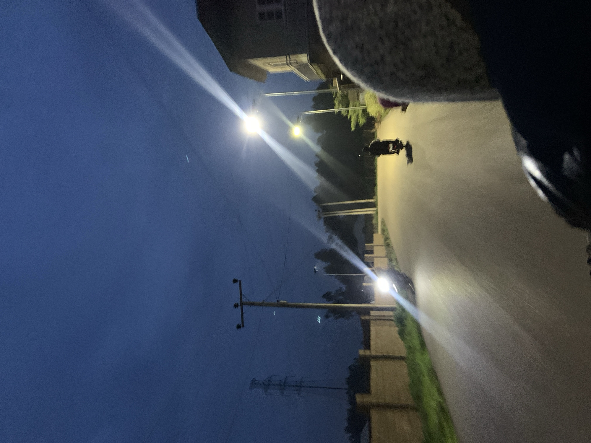 The image depicts a street scene at dusk or early night in Gura Top, Nigeria. The sky is a deep blue, transitioning from twilight to darkness, with a faint crescent moon visible in the upper left quadrant. The street is paved and wet, reflecting the light from street lamps and vehicle headlights.
In the foreground, a person's head and shoulder are visible, suggesting the image is taken from the perspective of someone riding on the back of a motorcycle. To the left, a light brown, stone-like wall runs along the side of the road, with patches of green grass growing beside it. Behind the wall, trees are silhouetted against the darkening sky.
Further down the road, a motorcycle with its headlight on is traveling away from the camera. The bright headlight illuminates the wet road surface, creating a strong beam of light. Another motorcycle, ridden by a person wearing dark clothing, is also moving away from the camera, its taillight faintly visible.
Several streetlights are visible, casting bright, elongated beams of light across the scene. One of these lights is particularly bright and positioned above and to the right of the road. In the background, a tall transmission tower stands against the sky. To the right of the road, a two-story building with windows is visible, illuminated by the streetlights. The overall atmosphere is that of a quiet evening commute on a suburban or semi-rural road.