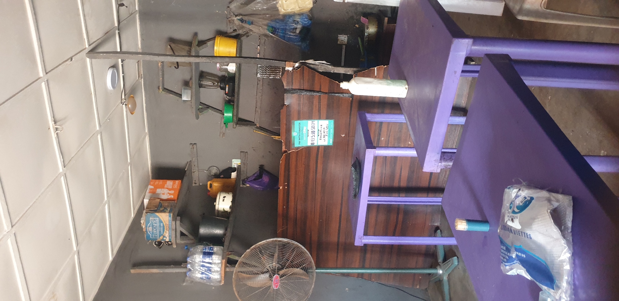 The image depicts a dimly lit interior of what appears to be a small shop or food stall. The walls are painted a dark gray, and the ceiling is made of white acoustic tiles. In the foreground, two purple tables are visible. On the left table, there's a plastic bag of what looks like napkins and a small cylinder with a toothpick. On the right table, a white plastic bottle stands upright.
Behind the tables, a wooden counter with a damaged top runs across the scene. Affixed to the counter is a green sign with payment information. To the left of the counter, a large, old-fashioned standing fan with a metal grill is positioned.
The background features several shelves mounted on the wall. The top shelf holds a blue box labeled "TOPCAFE COFFEE MIX" and an orange box with a white design. Below that, more shelves are sparsely populated with items including bottles of water, a metal pot, a yellow jug, a small white pot, and a purple plastic bag. Further to the right, another shelf holds a blender, green bowls, a pink container, and a yellow bucket. The overall impression is one of a functional, somewhat cluttered, and utilitarian space.
