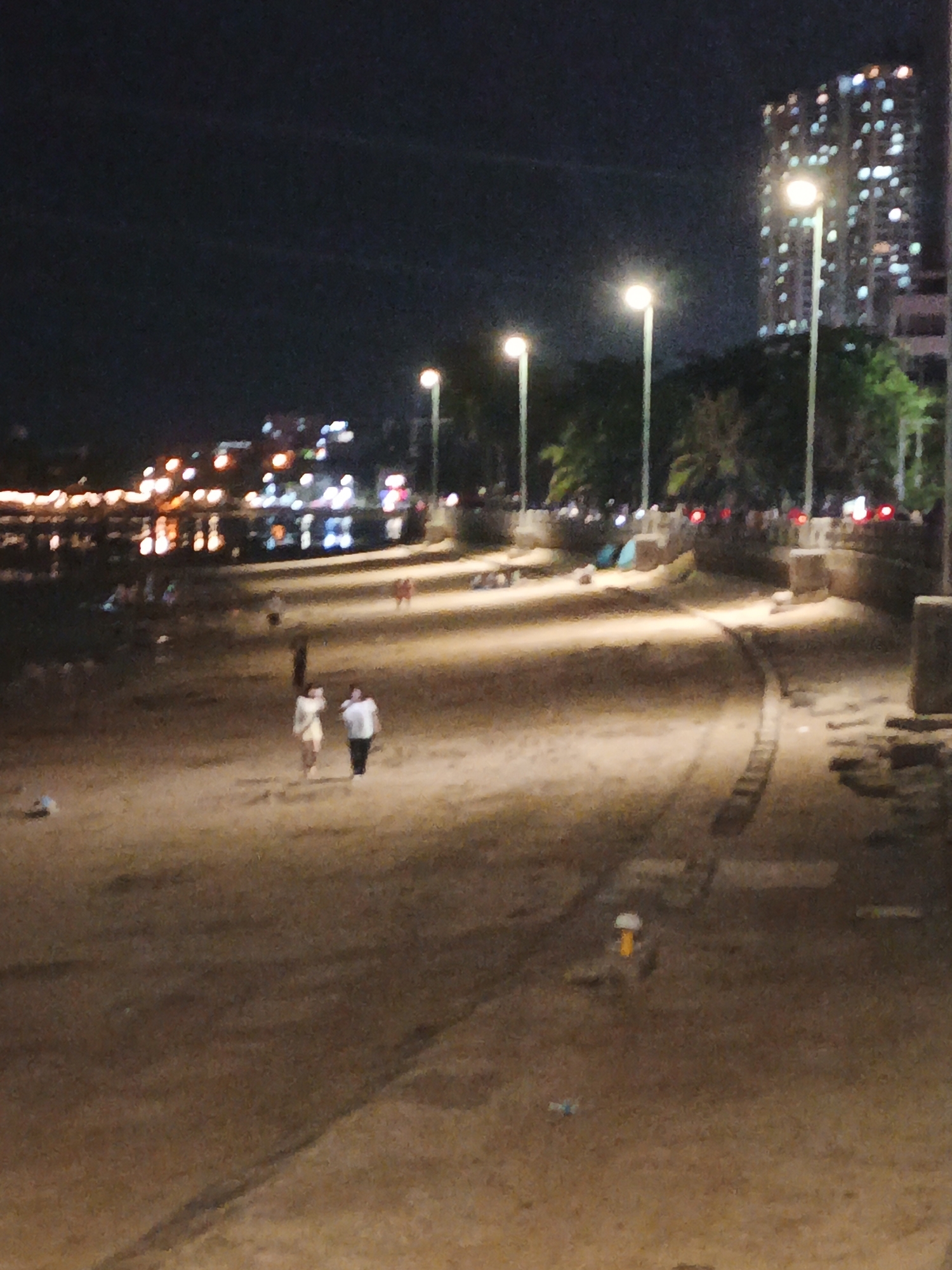 This is a nighttime photograph of a beach in Nha Trang, Vietnam. Two people are walking on the sand, illuminated by streetlights. In the background, a tall building with many lights is visible, along with more streetlights and palm trees. The water's edge, with its own lights, is in the distance, and there are several people scattered across the beach. The scene appears to be tranquil and public, likely a popular spot for evening strolls. The lighting suggests it is after dark, and the weather appears to be clear.