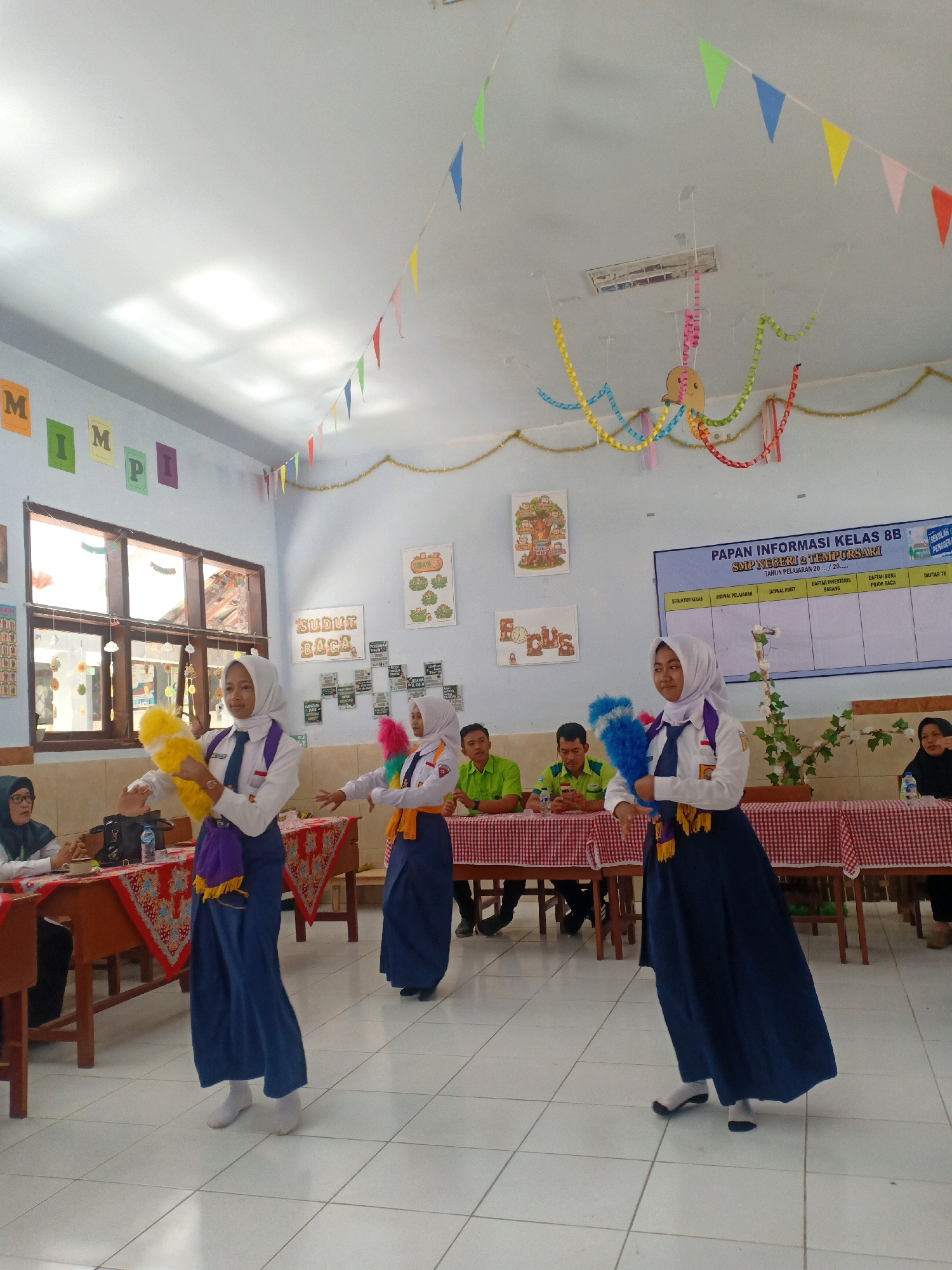 The image depicts an indoor scene, likely a classroom or an event space, decorated with colorful triangular flags and paper chains hanging from the ceiling. The room appears to be in Kaliuling, Indonesia, based on the provided context. In the foreground, three young women in school uniforms, consisting of white blouses, blue skirts, and headscarves, are performing a dance. Two of the dancers are holding props: one has a yellow and white fluffy prop in her right hand, and the other has a blue and pink fluffy prop in her left hand. Their movements suggest a choreographed performance.
Behind the dancers, several tables are set up with red and white checkered tablecloths. On these tables, there are water bottles and other items. Seated at these tables are adults, including men in green shirts and women wearing headscarves. Some are observing the performance, while others appear engaged in conversation.
The walls of the room are adorned with educational posters and handwritten notes. One poster on the right is a class information board for "KELAS 8B SMP NEGERI 2 TEMPURSARI". Another poster on the left wall has the word "MIMPI" written in large letters, with individual letters on separate colored squares. Other posters display text in Indonesian, such as "SUDI BACA" and "FOCUS". Large windows are visible on the left side of the room, allowing natural light to enter and casting shadows on the ceiling. The floor is tiled with white square tiles. The overall atmosphere suggests a school event or a cultural performance.