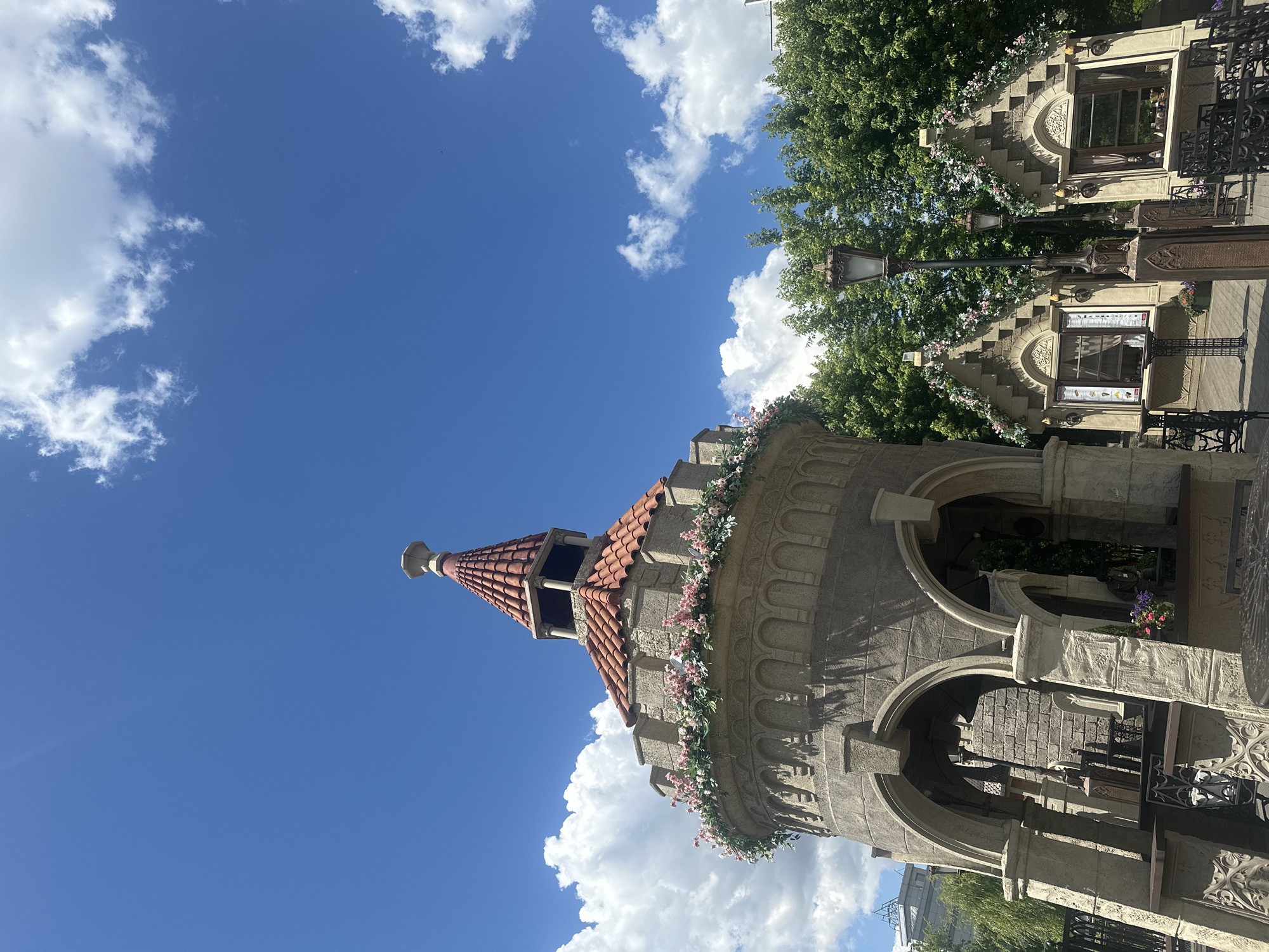 This media file depicts a whimsical, fairy-tale-inspired turret with a red tiled roof, set against a bright blue sky with scattered white clouds. The turret is adorned with floral garlands of pink and white flowers, adding to its charming aesthetic. To the right of the turret, a building with similar architectural elements can be seen, featuring arched windows and intricate stone detailing. A black wrought-iron fence partially obscures the building's facade, and a street lamp with an ornate design stands prominently in front of it. Lush green trees form a backdrop, suggesting a warm, sunny day. The scene evokes a sense of enchantment and possibly a tourist attraction or a themed establishment. There is no visible text in the image. The orientation suggests the photo was taken in Moscow, Russia.