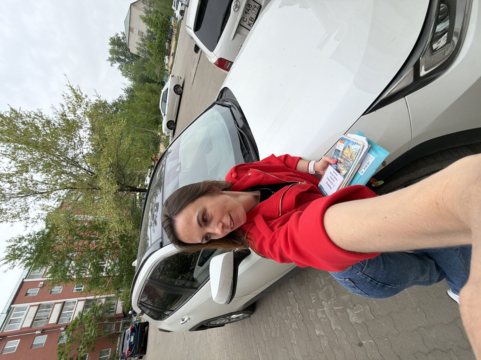 A woman is taking a selfie next to a white car in Birobidzhan, Russia. She is wearing a red jacket and jeans, and is holding some papers or brochures in her hand. The car is parked on a street with other cars and buildings in the background.