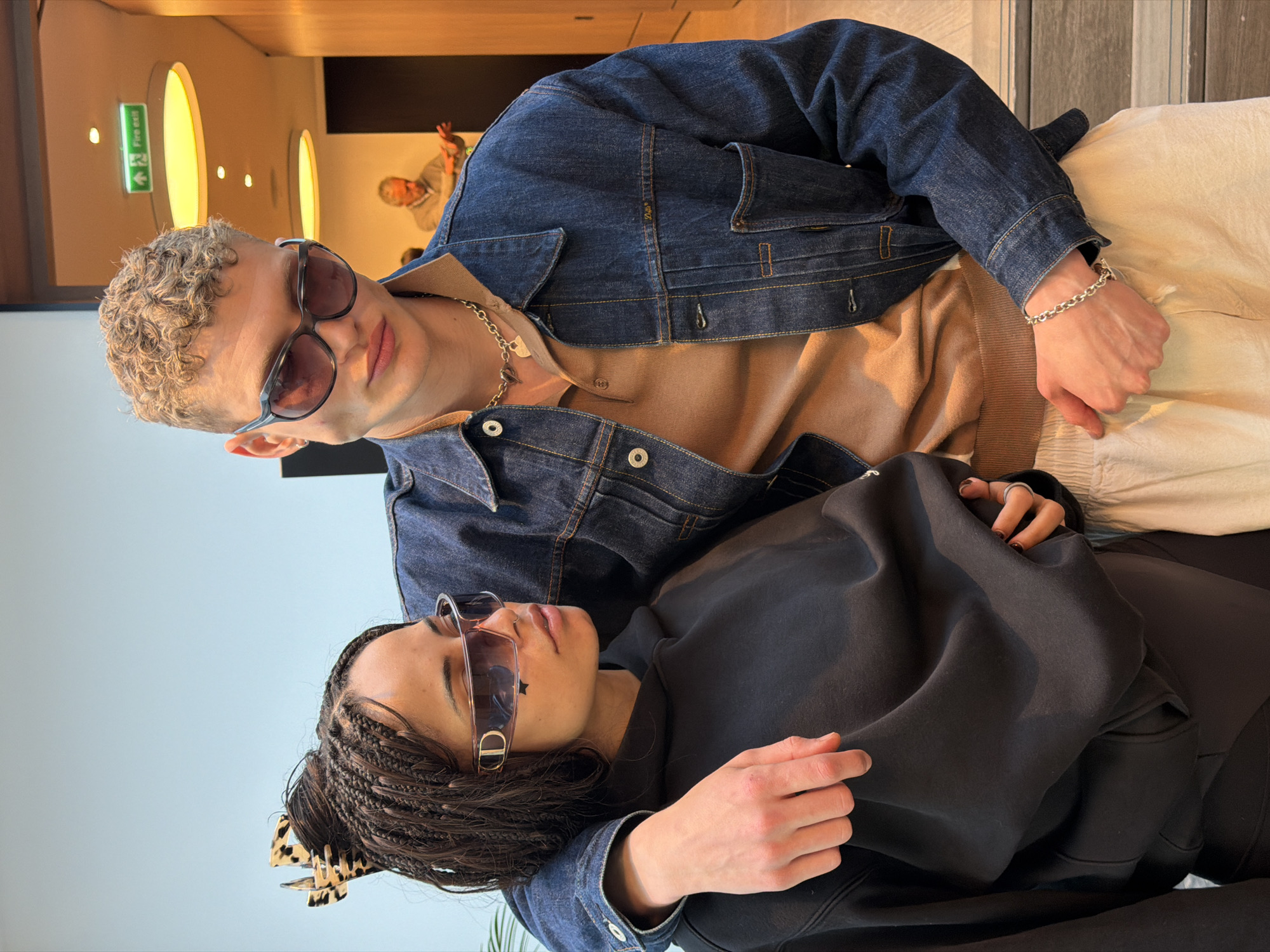 Two people are photographed indoors. The person on top is a light-skinned man with short, curly, blonde hair wearing sunglasses, a brown shirt, a blue denim jacket, a silver chain necklace, a silver bracelet, and cream-colored pants. The person underneath is a light-skinned woman with dark braided hair, wearing sunglasses, and a dark sweatshirt. The woman's left hand rests on the man's arm. The location shows a green "Fire Exit" sign with a person waving in the background. The location appears to be in London, United Kingdom.