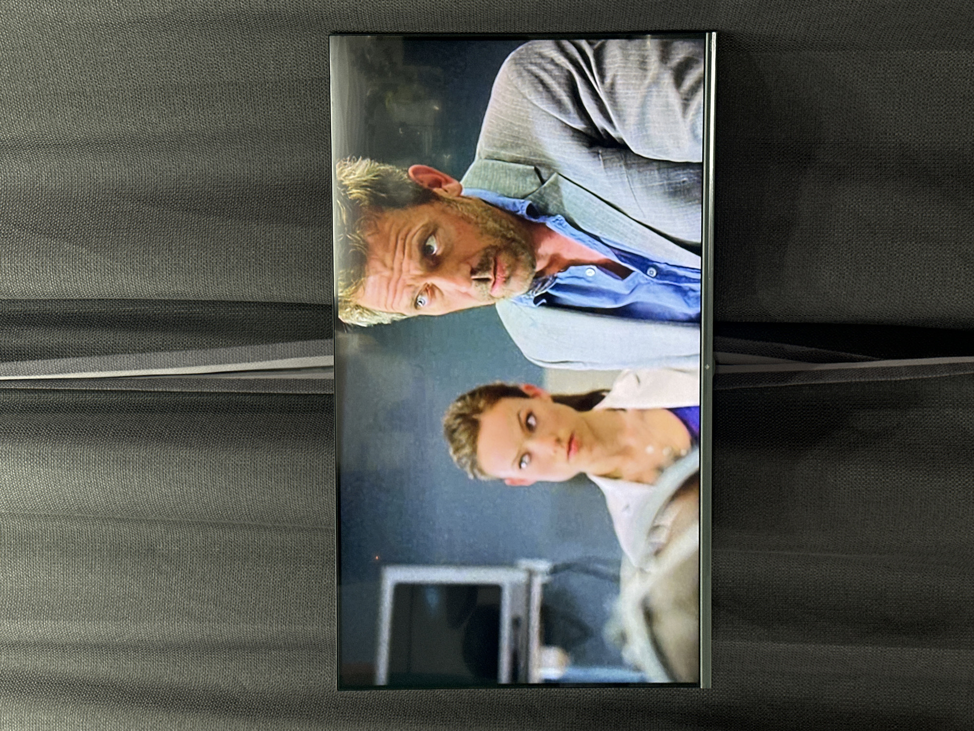A television screen displays a scene featuring actors Hugh Laurie and Jennifer Morrison from the TV show "House". The screen is situated against a background of a dark gray textured fabric. The location in Sochi, Russia, is not discernible from the image.