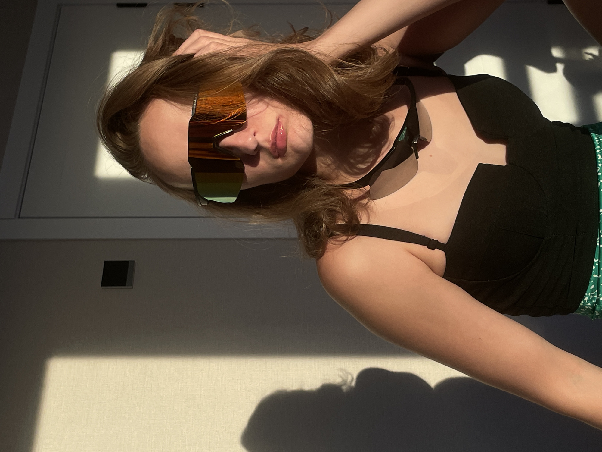 A young woman is indoors in Подшивалово, Russia, wearing sunglasses and a black top. She is lying down with one hand in her hair. Sunlight casts shadows on the wall behind her.