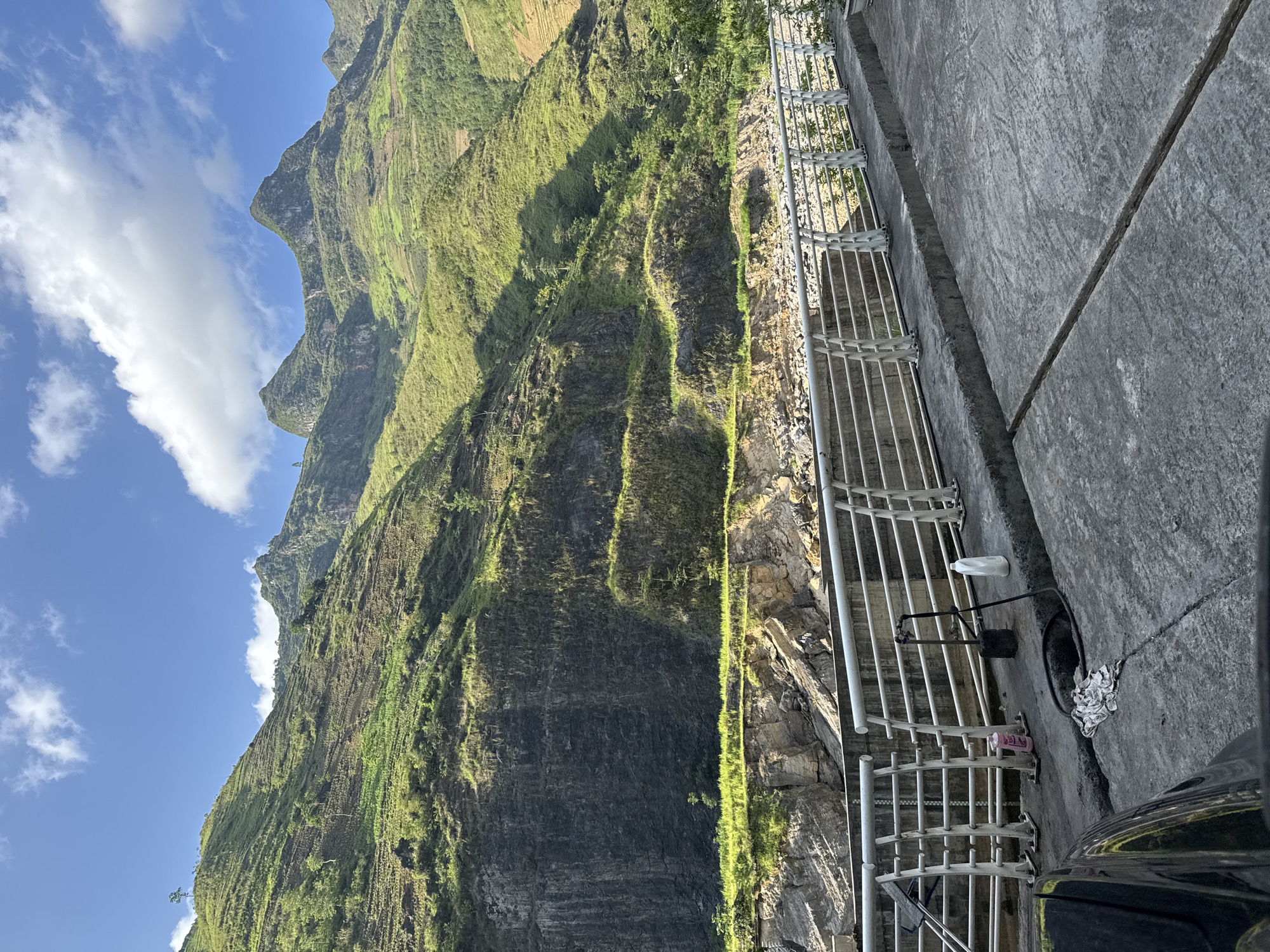 A scenic view in Mèo Vạc Commune, Vietnam, showcases a steep, verdant mountainside under a partly cloudy sky. Terraced formations are visible on the slope, and vegetation covers much of the terrain. A gray concrete wall and metal railing border the frame's right side, with some trash and debris lying on the concrete. A portion of a dark-colored vehicle is visible at the bottom.