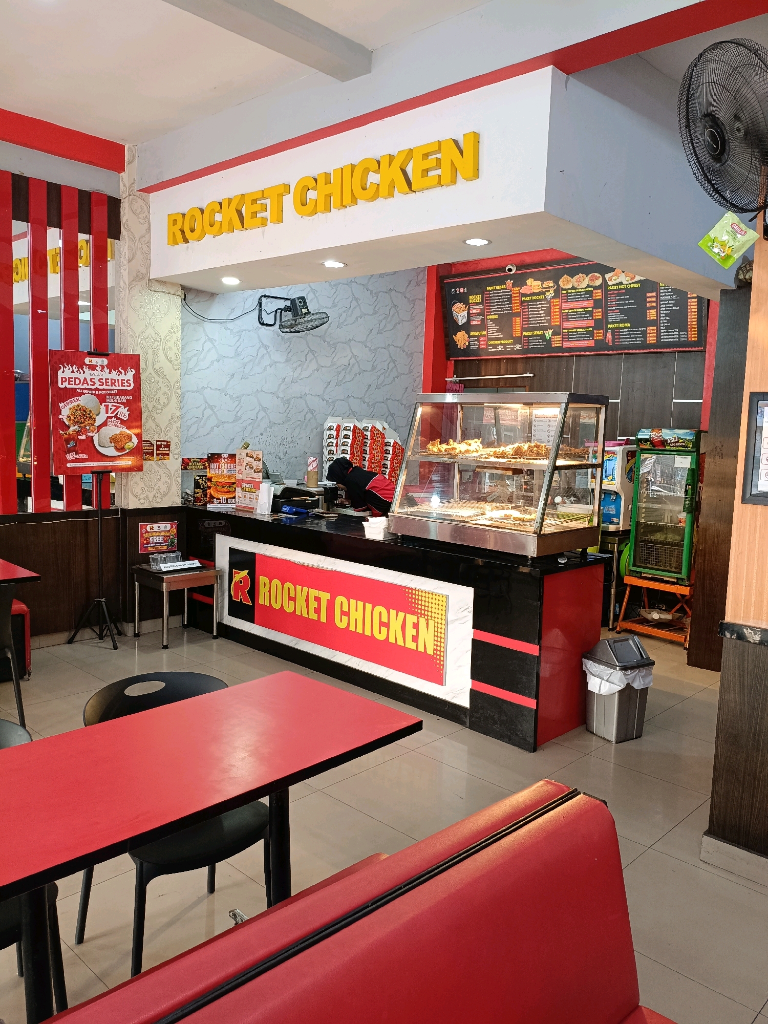 The image depicts the interior of a Rocket Chicken fast-food restaurant, likely located in Ngemplak, Indonesia, given the context provided. The establishment features a prominent yellow "ROCKET CHICKEN" sign above the counter area. Red and black are the dominant colors in the decor, seen on tables, seating, and the counter itself.
The counter area is staffed by at least one employee wearing a red and black uniform, with their face partially obscured by their head covering and the counter. They appear to be working behind the counter, potentially taking orders or preparing food. A display case filled with fried chicken is visible on the counter. A menu board with various food items and prices is mounted on the wall behind the counter.
In the foreground, red tables and red booth-style seating suggest a dining area for customers. A black fan is mounted on the wall, and a trash can is situated near the counter. Other elements visible include a refrigerator with a drink advertisement on it, a standee poster advertising a "PEDAS SERIES" (Spicy Series), and several boxes of what appear to be to-go containers stacked near the counter. The floor is tiled in a light color. The overall environment suggests a typical fast-food restaurant setting.