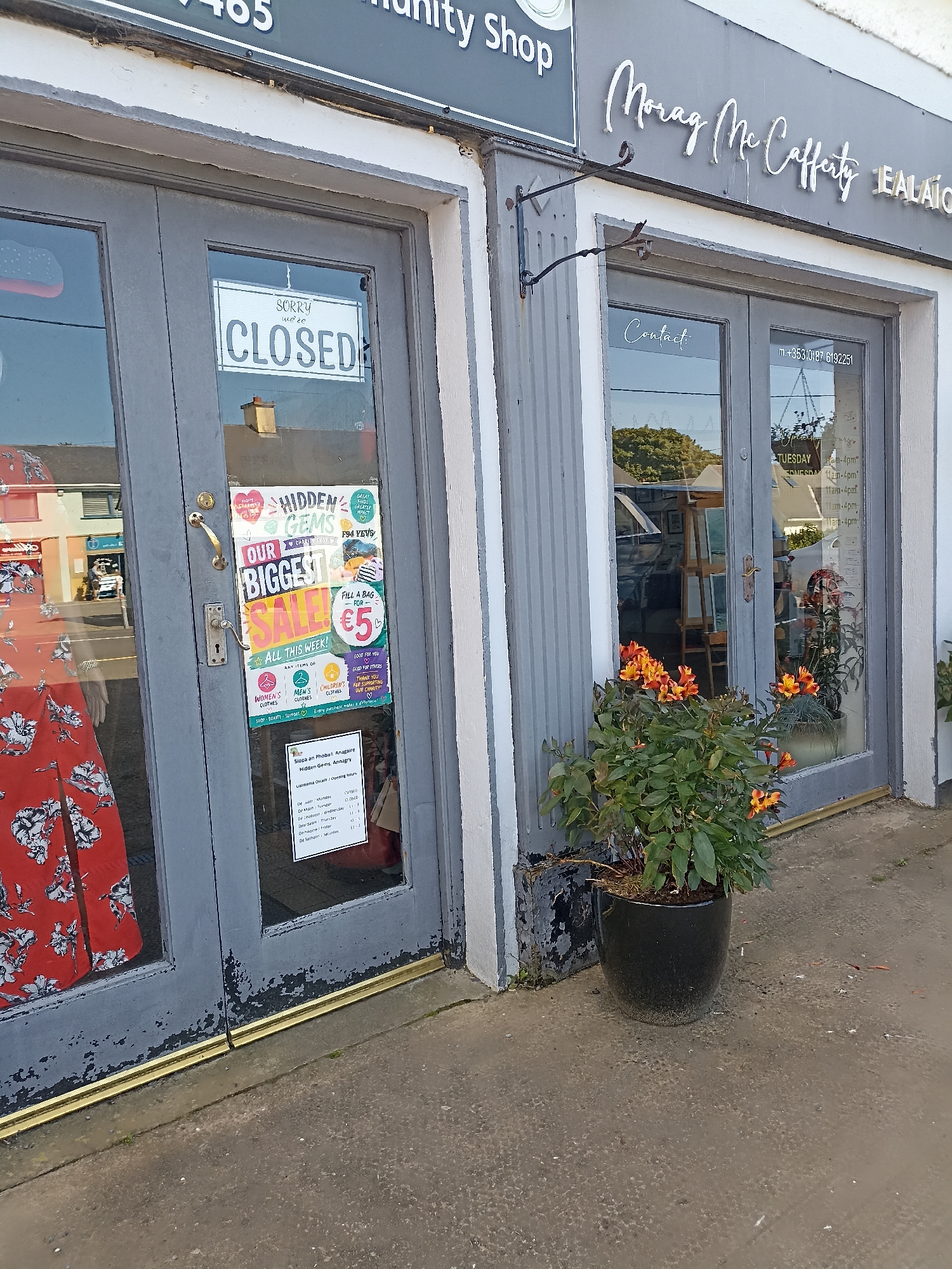 The image shows the exterior of a shop in Annagry, Ireland, with two gray double doors. A white sign reading "SORRY we're CLOSED" is affixed to the left door. On the same door, a colorful poster advertises a "BIGGEST SALE" for "ALL THIS WEEK!" and "ANY ITEMS OF CLOTHING" for "€5". Below this, icons representing "WOMEN'S CLOTHES", "MEN'S CLOTHES", and "CHILDREN'S CLOTHES" are displayed. Further down, a printed schedule details "Opening Hours" for "Hidden Gems Annagry", listing opening times from Monday to Saturday.
The right door has a sign with "Contact" written in cursive. A phone number, "m.+353(0)87 6192251", is also visible. Inside the shop, through the glass, shelves and various items are faintly discernible. A potted plant with orange and yellow flowers sits in front of the right door. The shop's facade features the name "Morag McCafferty" in prominent white lettering. The environment appears to be outdoors, with a concrete sidewalk in the foreground and a building visible across the street in the reflection of the left door. The overall condition of the doors shows some peeling paint.