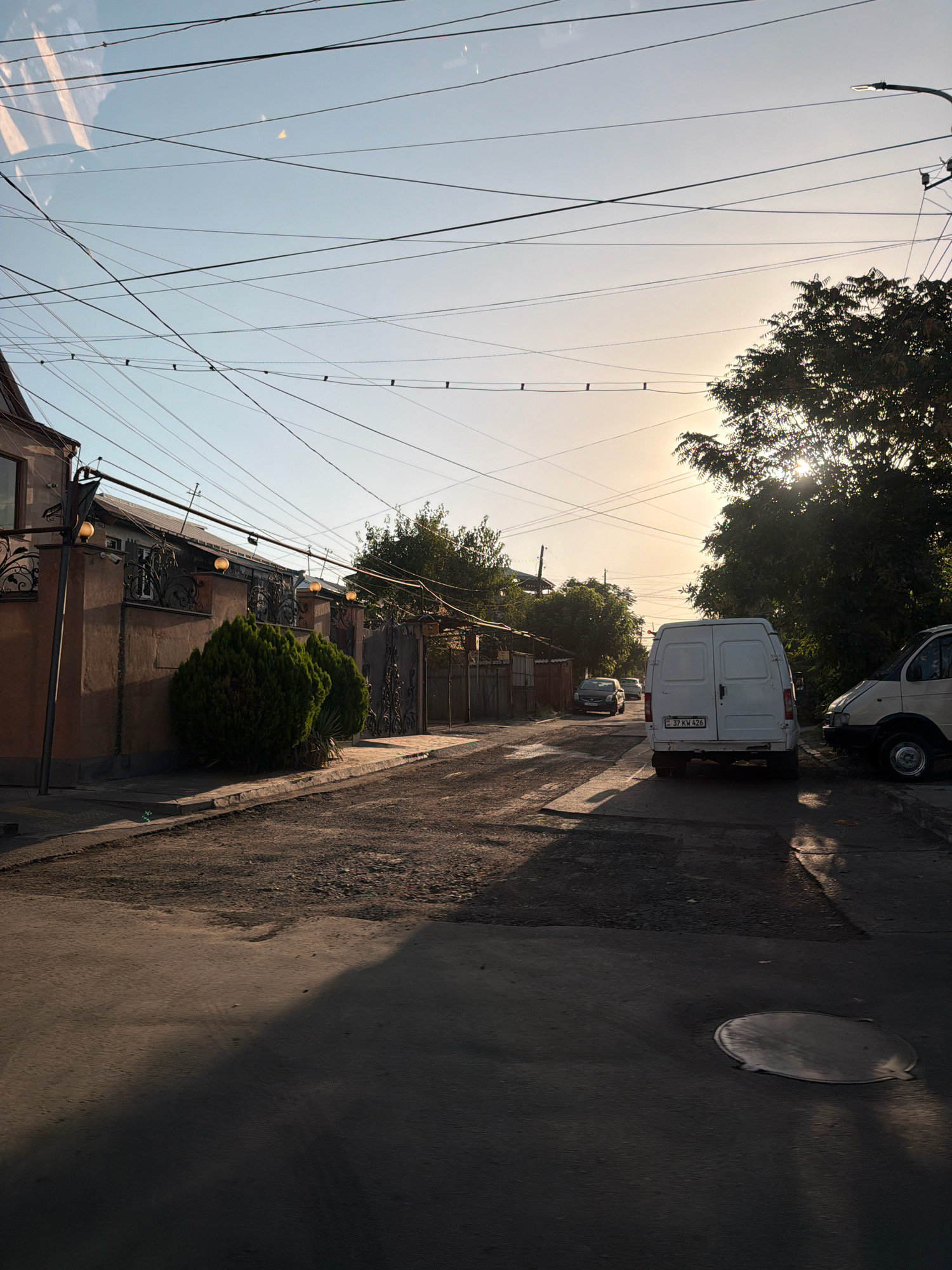 The image depicts a street scene in Yerevan, Armenia, likely during the late afternoon or early evening, given the low angle of the sun casting long shadows. The sky is clear with a gentle gradient of pale blue and yellow, indicative of sunset or sunrise. Overhead, a complex network of electrical wires crisscrosses the frame, a common sight in urban Armenian landscapes.
On the left side of the street, a traditional Armenian house is visible, characterized by its light brown stucco facade, ornate metal fencing, and decorative light fixtures. Two neatly trimmed evergreen bushes are positioned in front of the house, adding a touch of greenery.
The street itself is paved with asphalt, showing signs of wear and tear, with patches of gravel and dirt. Several vehicles are present: a white cargo van is parked on the right side of the road, displaying Armenian license plates (37 KV 426). A lighter-colored van is parked partially behind it. Further down the street, two cars are driving away from the camera, one a dark sedan and the other a lighter-colored hatchback.
The right side of the street is lined with trees and a fence, casting shadows that stretch across the road. The overall impression is of a quiet, residential street in a city undergoing its daily rhythm. The scene is devoid of any visible people.