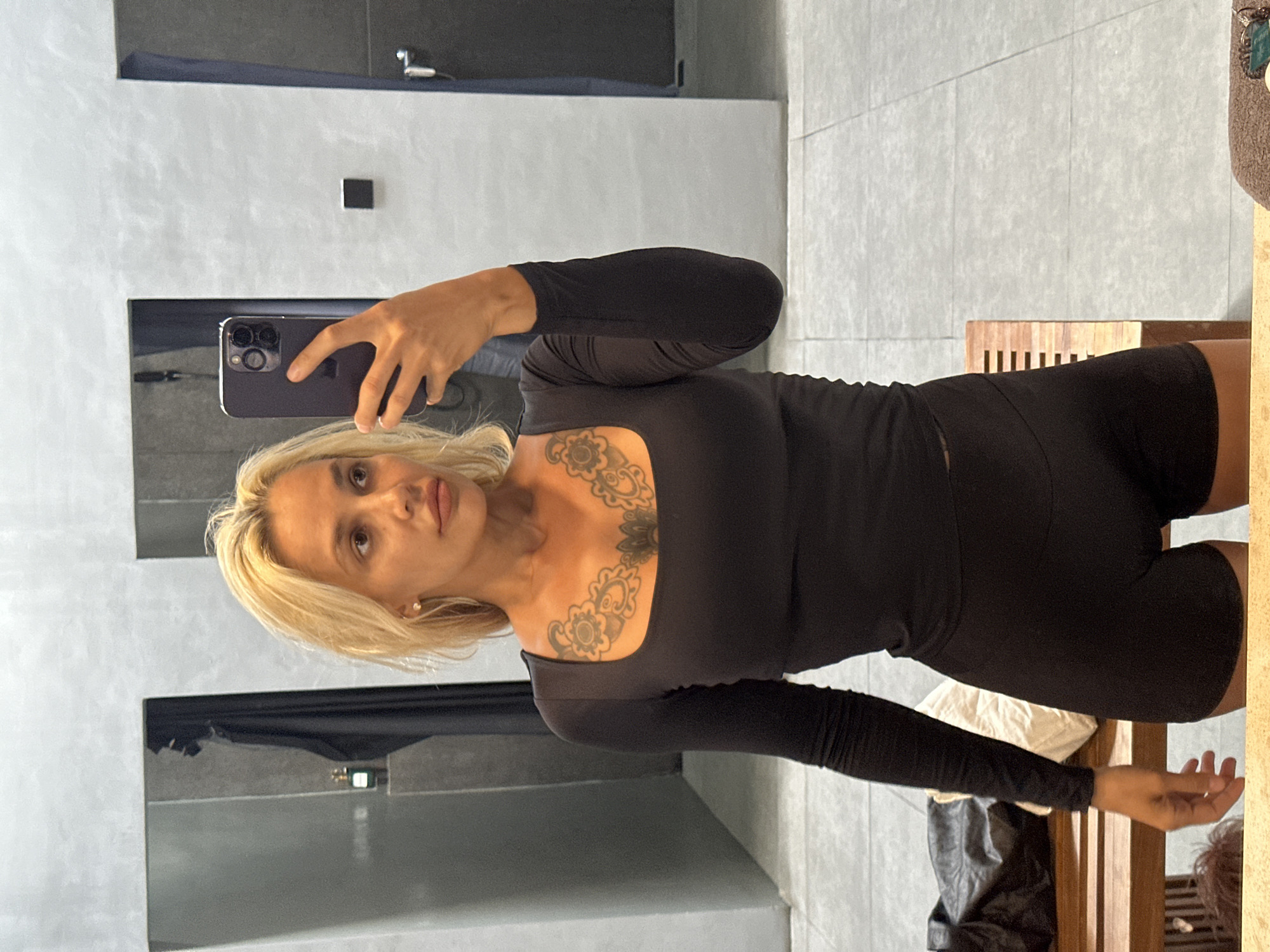 A blonde woman with tattoos on her chest is taking a selfie in a locker room in Kuta Selatan, Indonesia. She is wearing a black long-sleeved top and black shorts. She is holding a dark-colored smartphone in her right hand. In the background, there are lockers and a wooden bench. The floor is tiled.