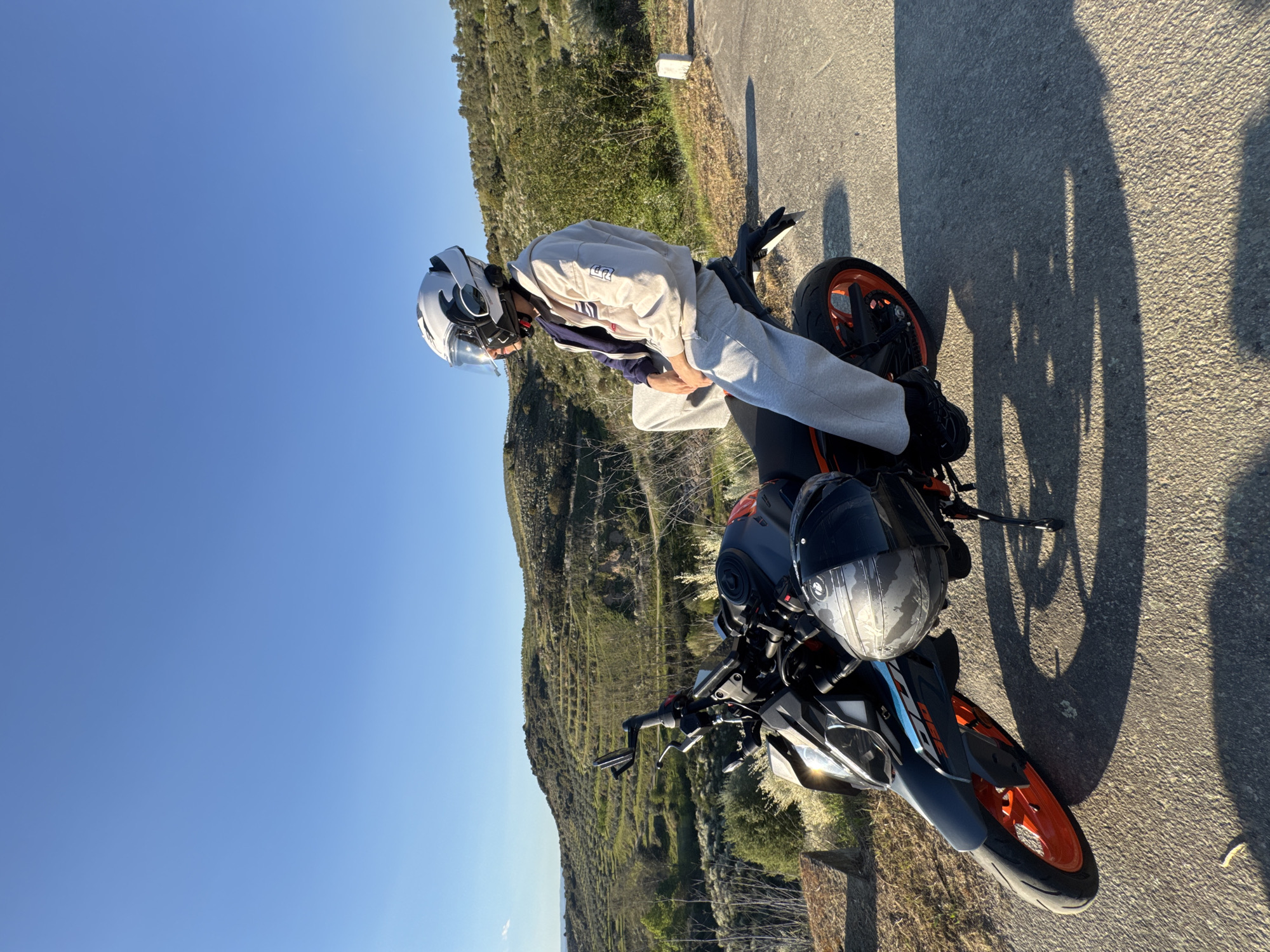 A person wearing a white helmet and grey clothing is seated on a black and orange motorcycle parked on an asphalt road. Another helmet, black, rests on the front of the motorcycle. The background consists of a hillside with vegetation and a blue sky. The location is Mirandela, Portugal.