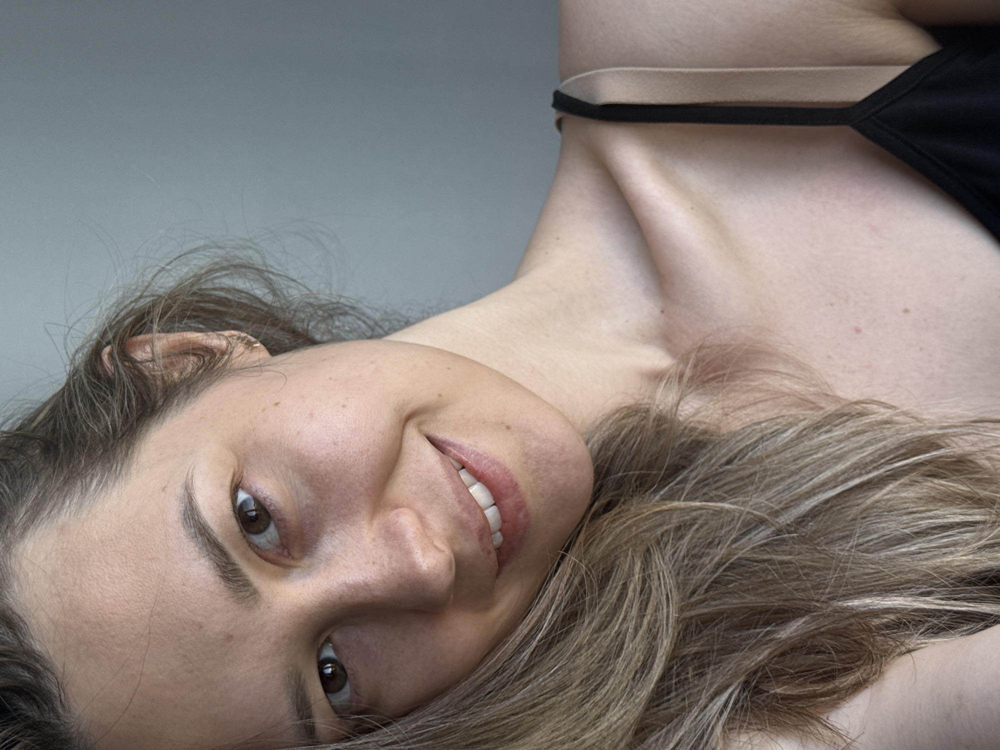 A close-up, angled portrait depicts a woman with light skin and brown hair, smiling. Her head is tilted to the left. She is wearing a black top with a tan strap visible. The background is a neutral grey. There is no indication of the city of Chaikovsky, Russia, or any specific location information.