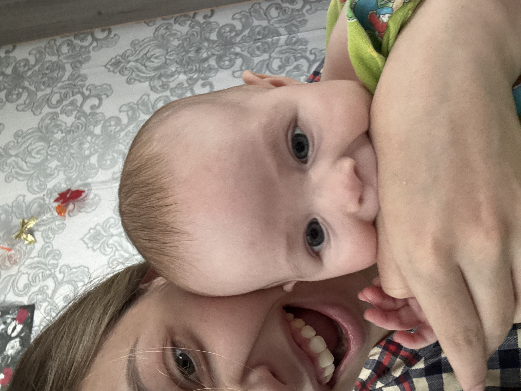 A rotated close-up shot depicts a baby lying on top of a woman, with both faces in view. The baby has blue eyes and appears to be biting the woman's hand. The woman has brown hair and is smiling widely, showing her teeth. The background includes a patterned wall and other indistinct objects. The location is Кемуль, Russia.