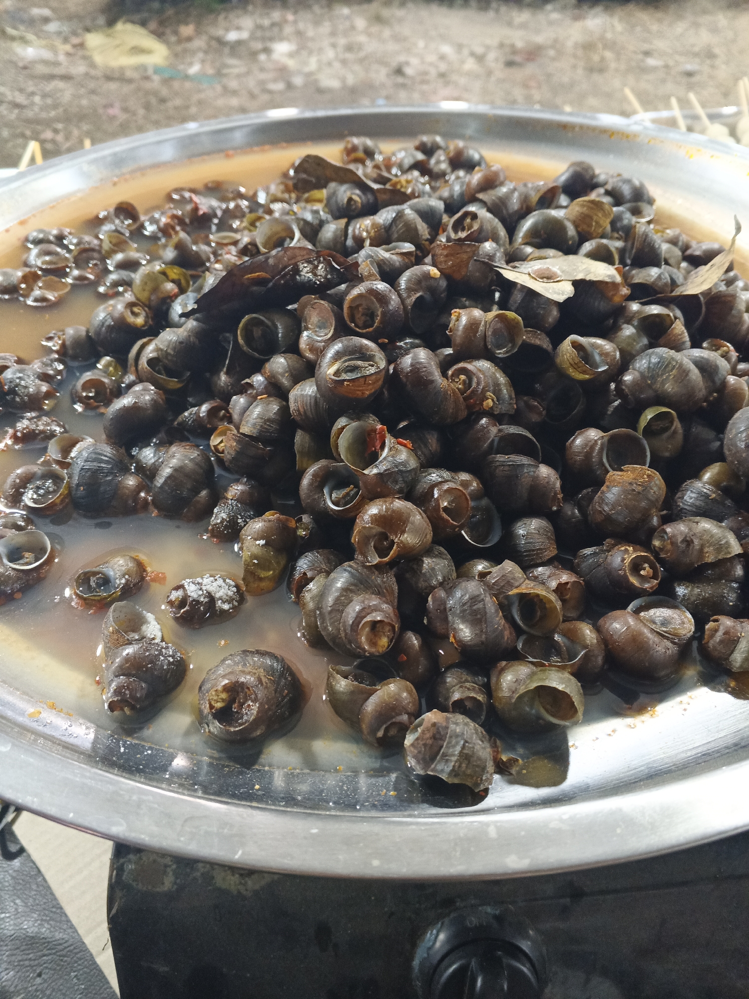 A shallow, wide, silver metal platter is filled with a dense pile of small, dark brown to black, spiral-shelled snails. The snails are submerged in a reddish-brown liquid, which appears to be a broth or sauce. Some of the snails have their openings facing upwards, revealing the interior of their shells. A few dry, brown leaves are scattered amongst the snails. The platter rests on what appears to be a black, textured surface, possibly a stove or a cooking apparatus, with a black knob visible in the lower right corner. The background is blurred and suggests an outdoor or semi-outdoor setting with a dirt or gravel ground and some indistinct objects. The lighting is somewhat dim, casting soft shadows. The image appears to be taken at a street food stall in Sukabumi, Indonesia, likely during a preparation or serving of a local delicacy. There are no people or animals directly visible in the image.