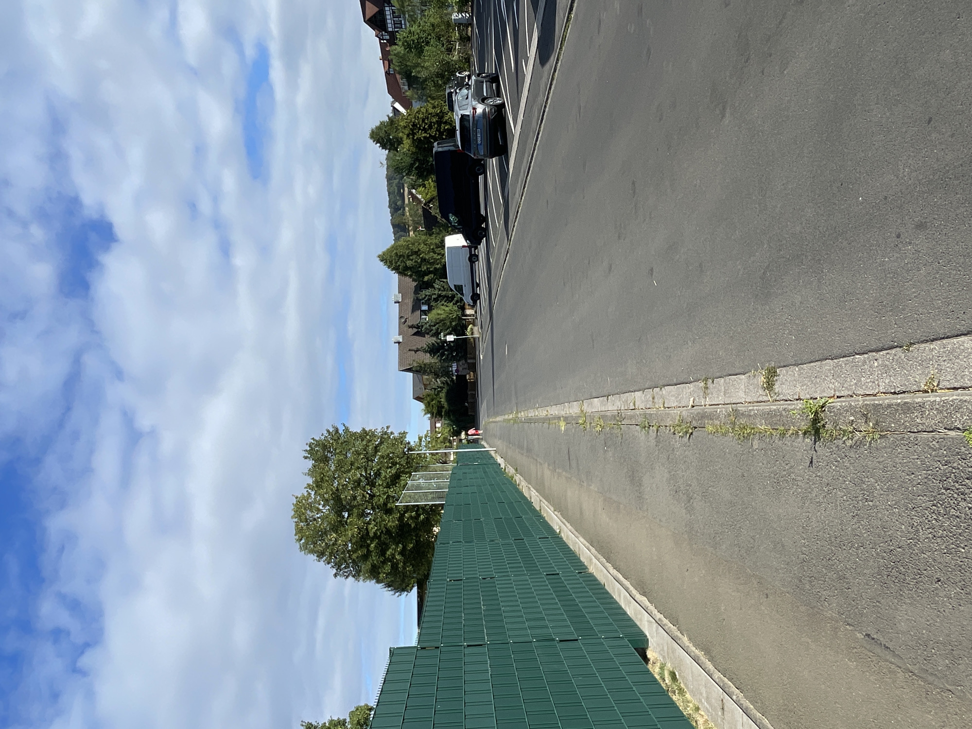 The image depicts a street scene in Neukirchen, Germany, on a bright, partly cloudy day. A long, green fence runs along the left side of the frame, bordering a paved sidewalk. Beyond the sidewalk, a two-lane asphalt road extends into the distance.
On the road, several vehicles are visible. A dark SUV is parked on the right side, facing away from the camera. Further down the road, a black van and a white van are parked. In the background, houses with pitched roofs, typical of German architecture, are nestled among mature trees. The sky is a vibrant blue, dotted with fluffy white clouds.
Two individuals, appearing to be adults, are walking away from the camera on the sidewalk in the distance. They are dressed in casual clothing. The overall scene suggests a peaceful residential area in Neukirchen.
