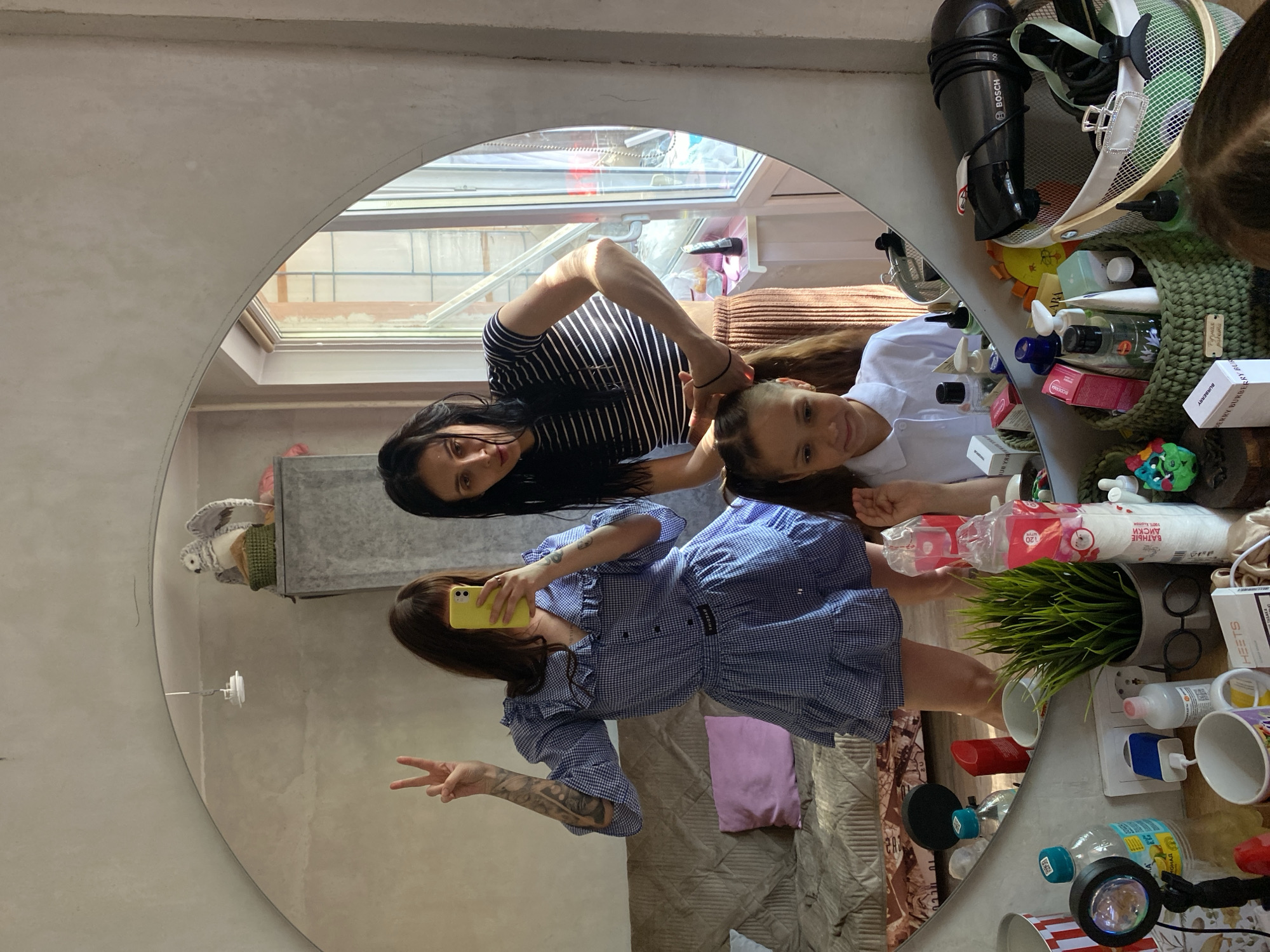 A photograph depicts two women posing in front of a large, circular mirror. One woman is wearing a blue and white checkered dress with frills, and she is holding a yellow cell phone in her hand, presumably taking the picture. She is making a peace sign with her fingers. The other woman, positioned closer to the top of the mirror, is wearing a black and white striped shirt and appears to be styling or touching the other woman's hair.
The mirror reflects an interior space, including a window with a view of a building exterior and a cluttered table or shelf area. Various objects are visible on the surface, including toiletries, cosmetics, a potted plant, water bottles, and other miscellaneous items. The overall setting appears to be a domestic or personal space.
There is no mention of the city Votkinsk, Russia, in the image, so I cannot include that information in the description.